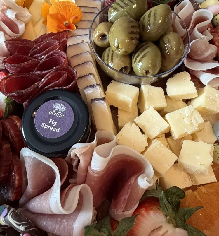 A cheese and charcuterie board with green olives in a glass bowl, cubes of cheese, slices of cured meat, a jar of fig spread, and strawberries, garnished with an orange flower.