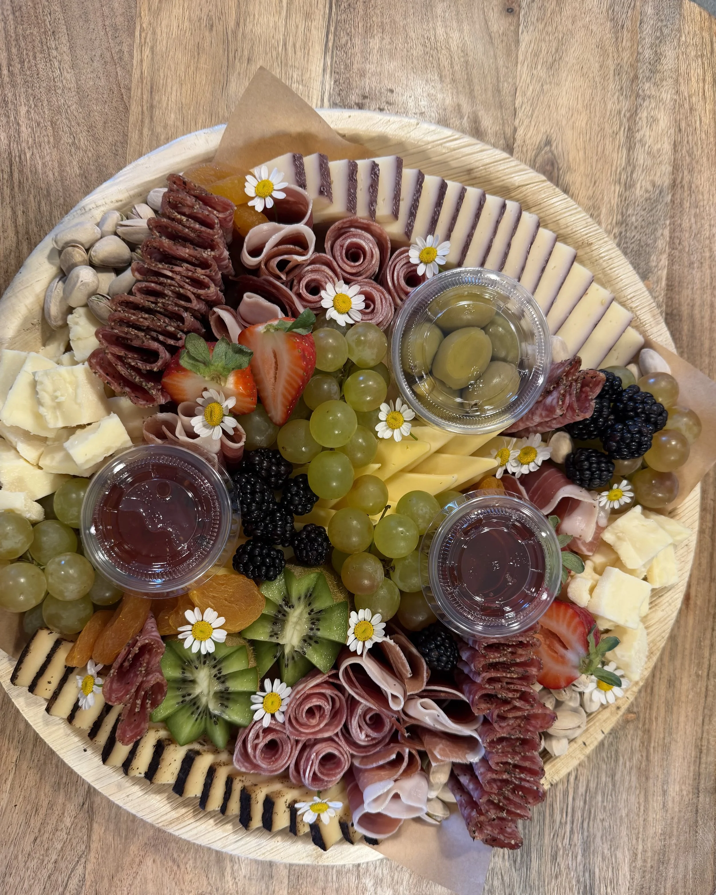 A large round wooden platter with assorted cheeses, cured meats, fresh fruit, and small flowers. There are slices of cheese with blackened edges, rolled cured meats, green grapes, strawberries, blackberries, kiwi, orange slices, and small white daisies used as decoration. Small containers with sauces or dips are also included.
