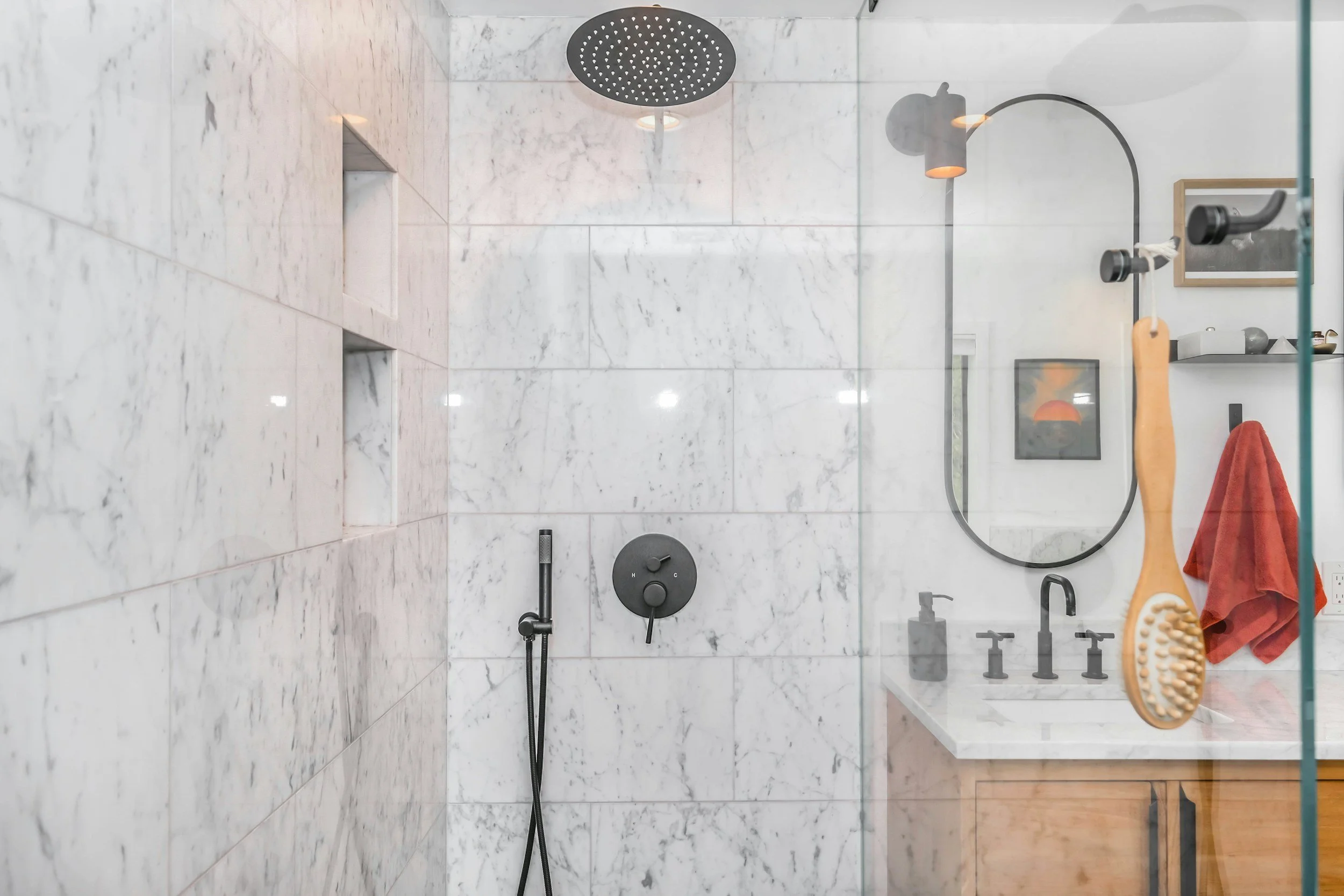 Modern bathroom with white marble walls, black showerhead, handheld shower, mirror, red towel, wooden hairbrush, and framed art on the wall.
