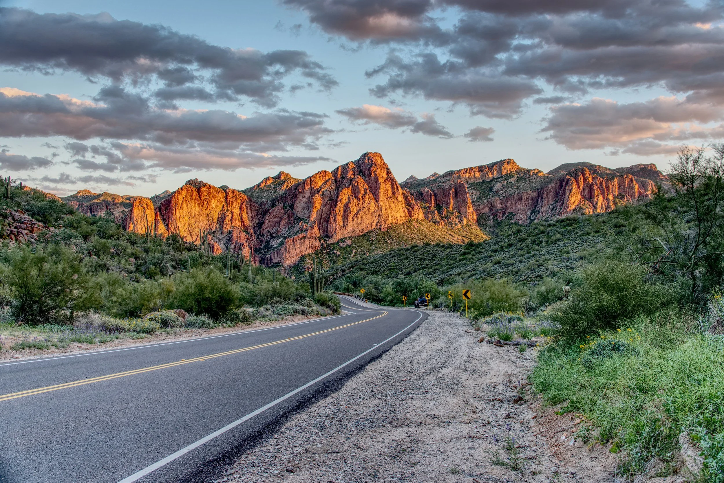 A winding road through a desert landscape with rocky mountains in the background under a partly cloudy sky, during sunset.
