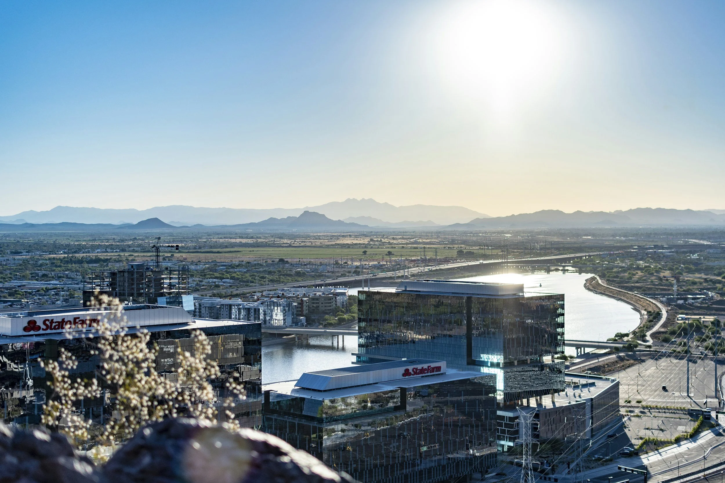 A cityscape with modern buildings, a waterway, and mountain range in the distance under a clear sky with the sun shining.