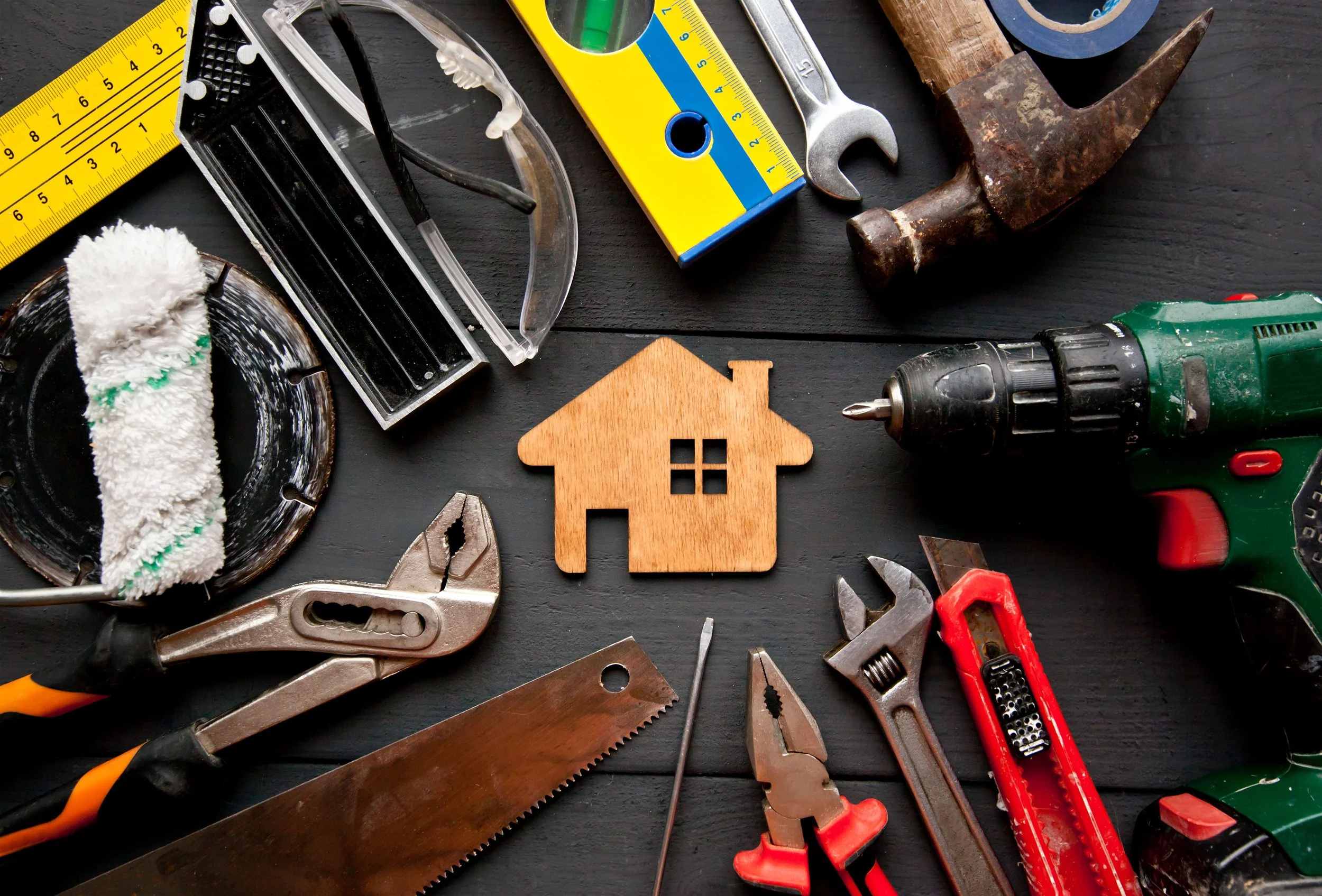 Handyman tools, including hammer, drill, saw, wrench, eye protection, pliers, level, screwdriver, and blade. Wooden home cutout in center of image.