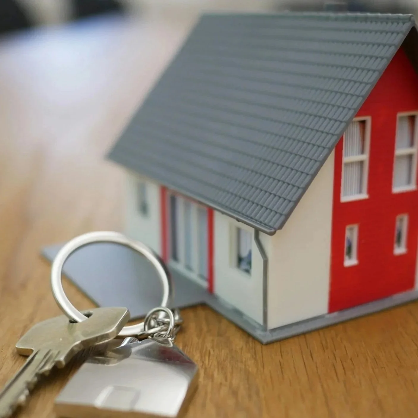 A small model house with a red and white exterior, a gray tiled roof, and multiple windows. Keys and a keyring are placed on a wooden surface in the foreground.