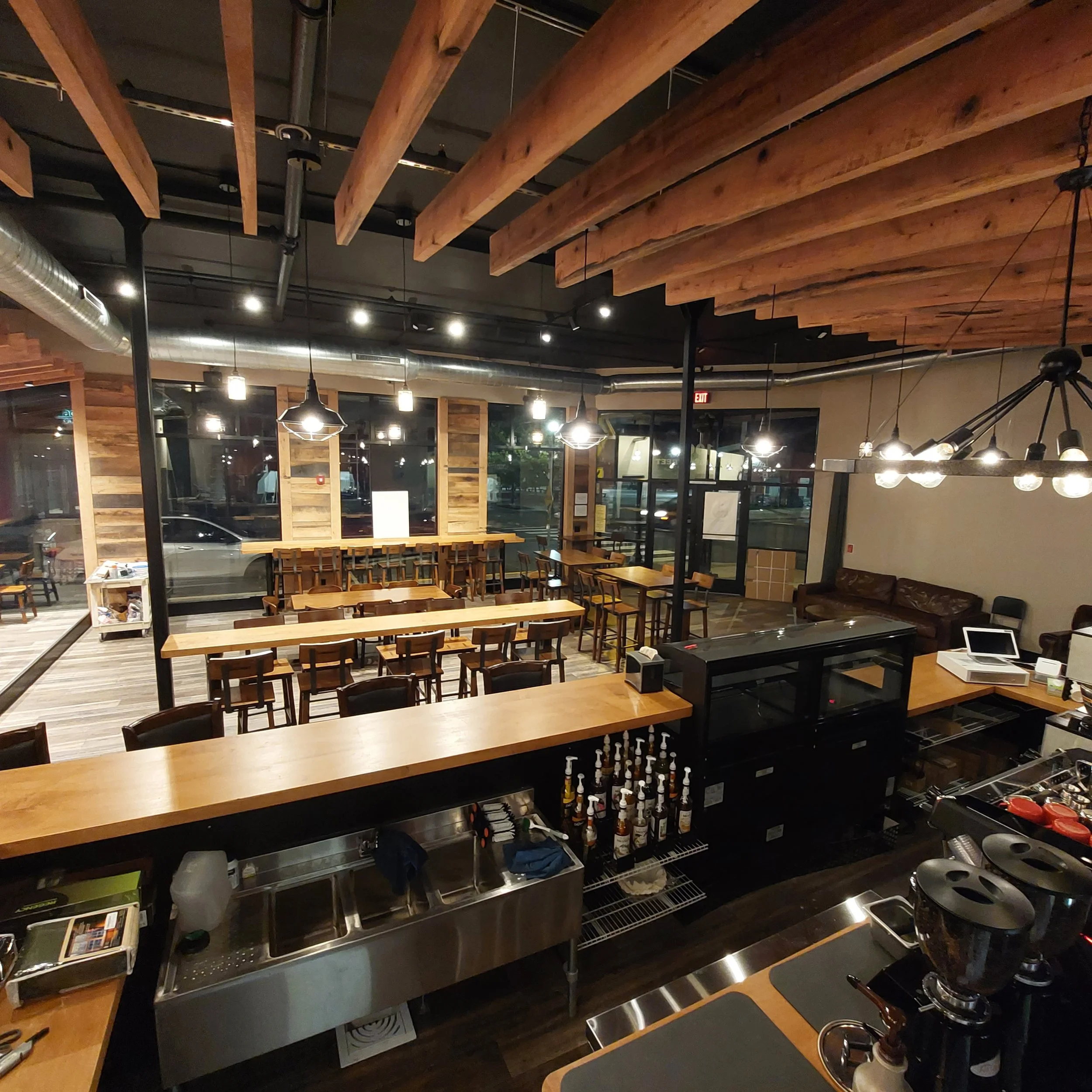 Interior of a modern coffee shop or cafe with wooden beams on the ceiling, pendant lights, and large windows. There are tables and chairs arranged for customers, a counter with various bottles and equipment, and lounge seating.
