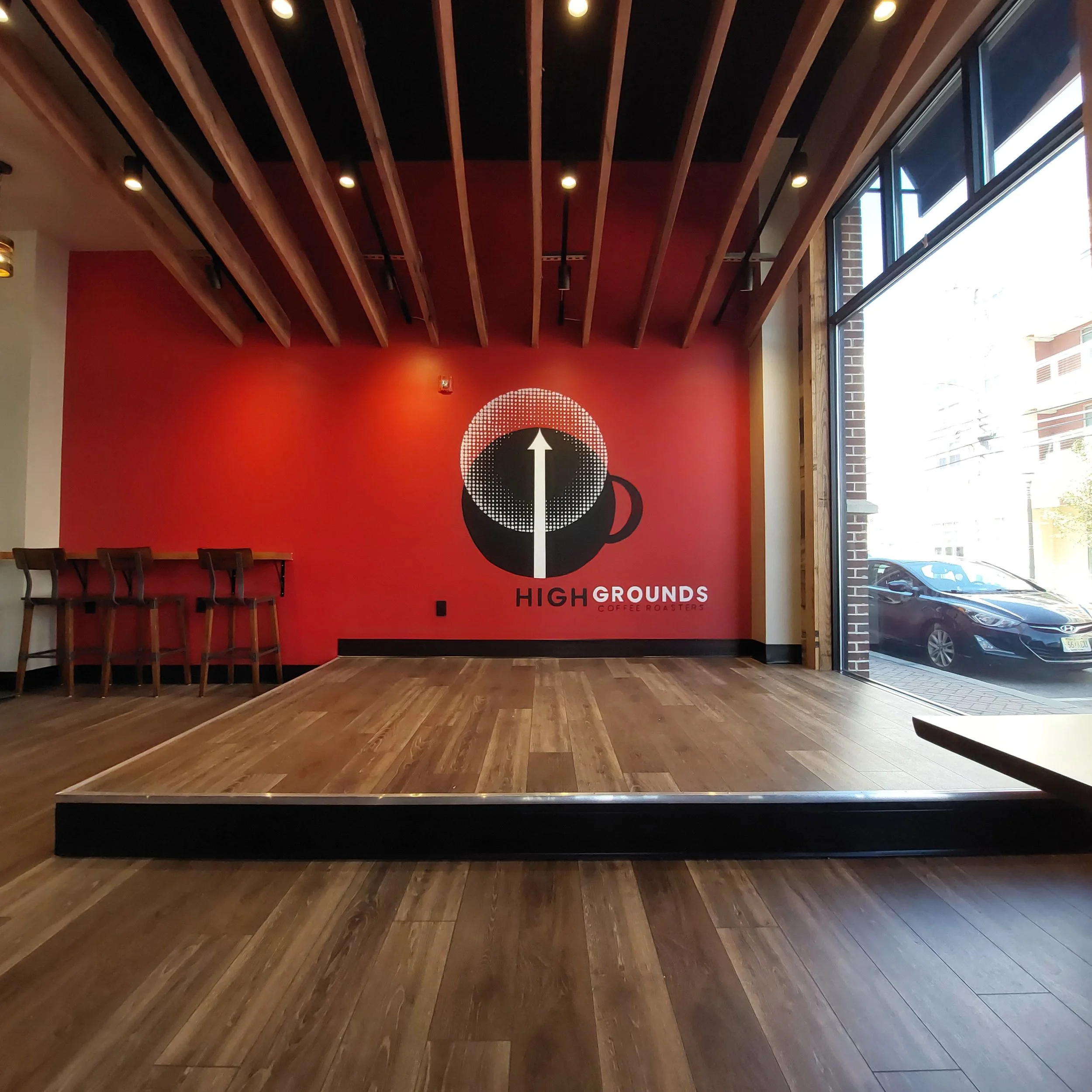 Interior of a coffee shop with a red wall featuring a logo of a coffee cup with an upward arrow, the words 'High Grounds Coffee Roasters', and a small stage area with wooden floors and a bar counter with chairs on the left side. Large windows on the right allow natural light in.