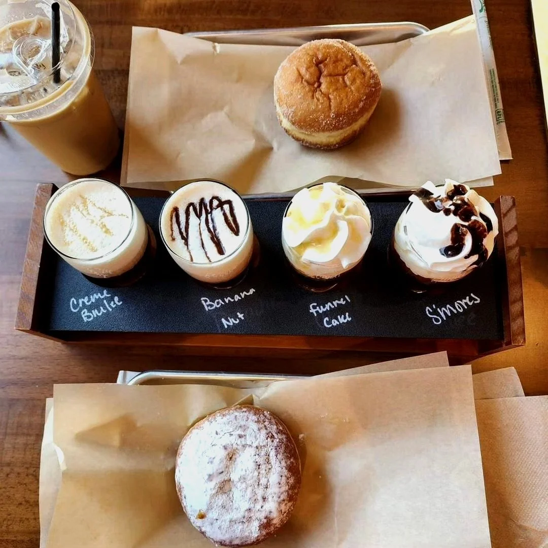A table with two desserts: a muffin with powdered sugar and a tray of four shot glasses with different dessert flavors labeled Creme Brulee, Banana Nut, Fannie Call, and S'mores, along with a tall iced coffee.