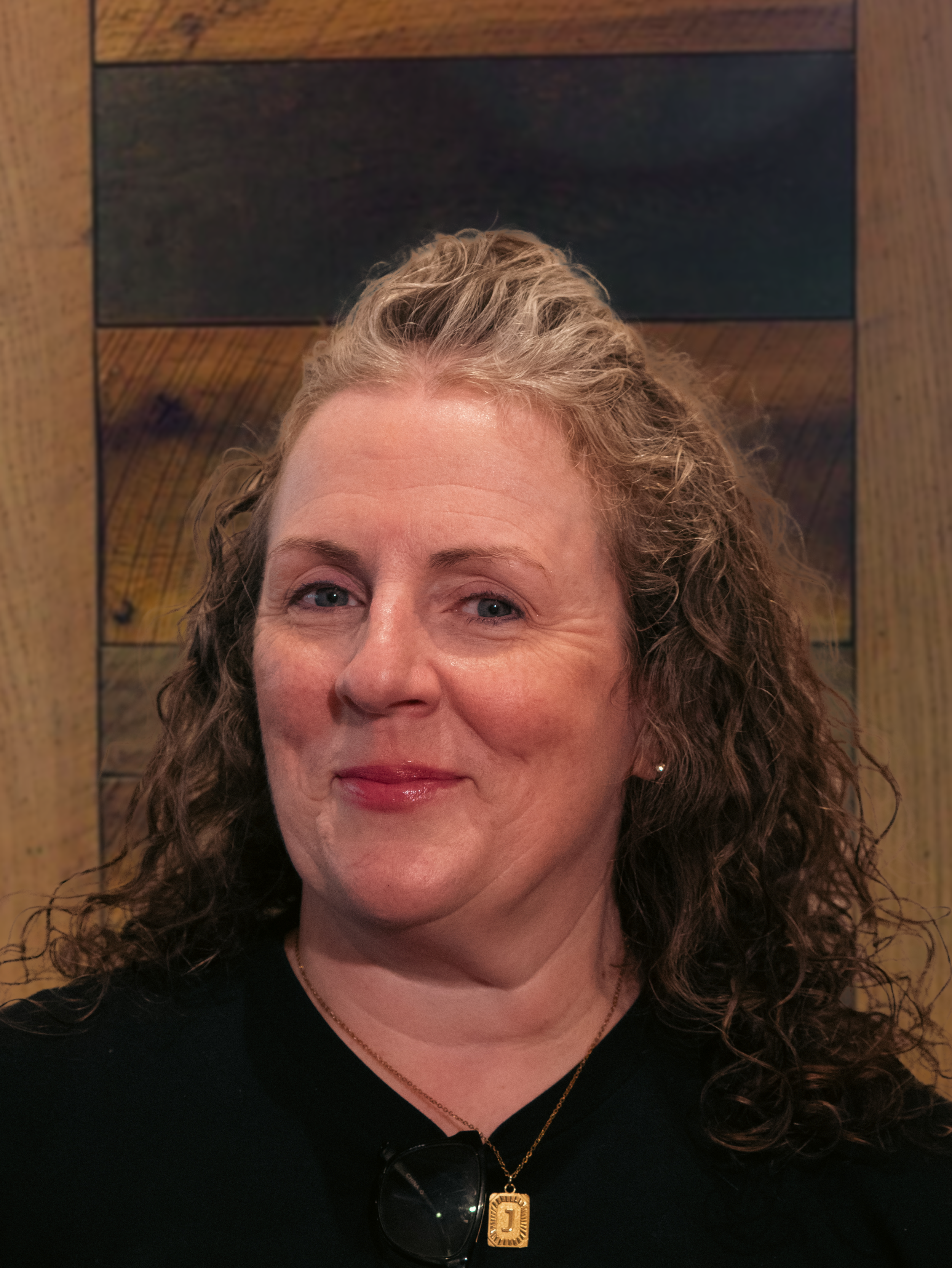 A woman with curly blonde hair smiling, wearing a black top, sunglasses hanging from her collar, and a gold necklace with a pendant, in front of a wooden background.