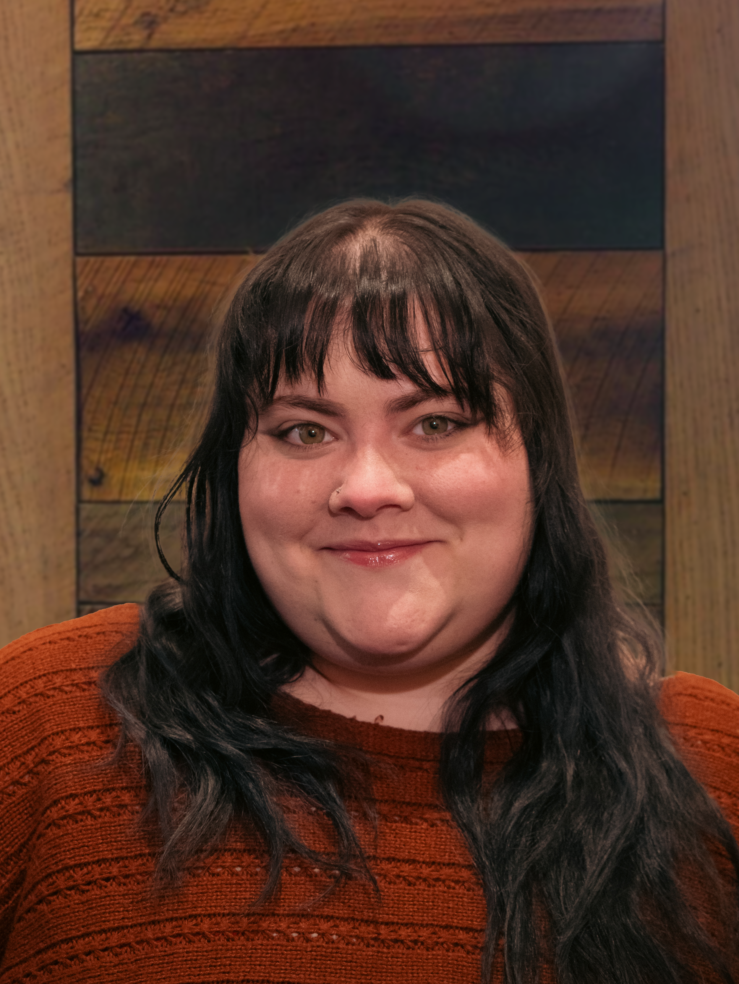 A woman with long dark hair and bangs, smiling at the camera, wearing an orange sweater, against a wooden background.