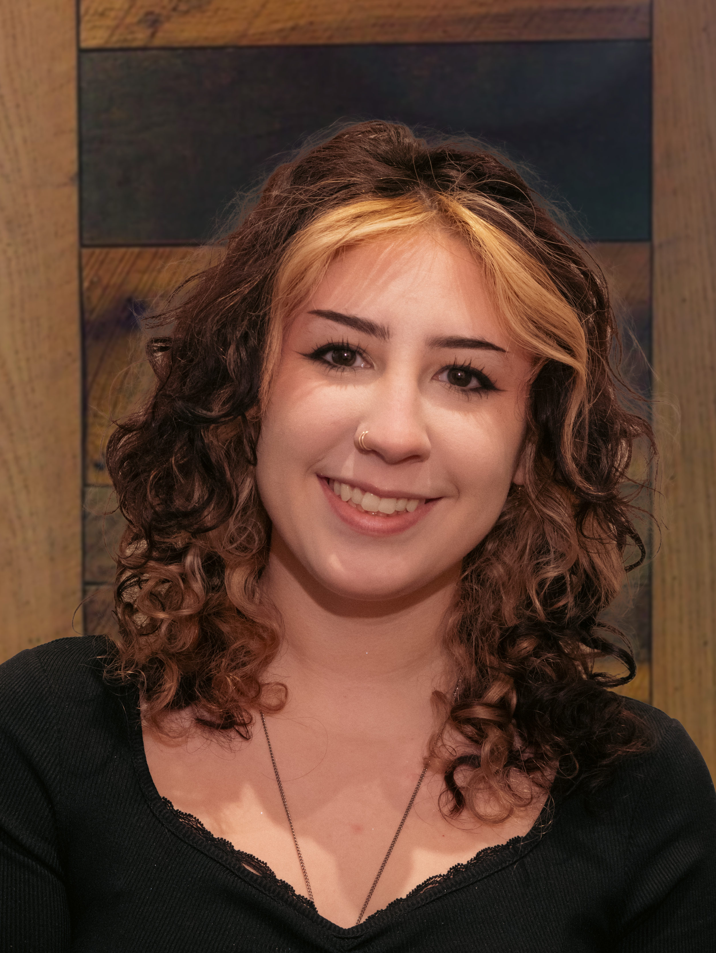 A young woman with wavy, shoulder-length hair that is dark brown with blonde highlights on top, smiling at the camera. She has a nose ring, wears a black top with a lace neckline, and a thin necklace. Behind her is a wooden wall or panel.