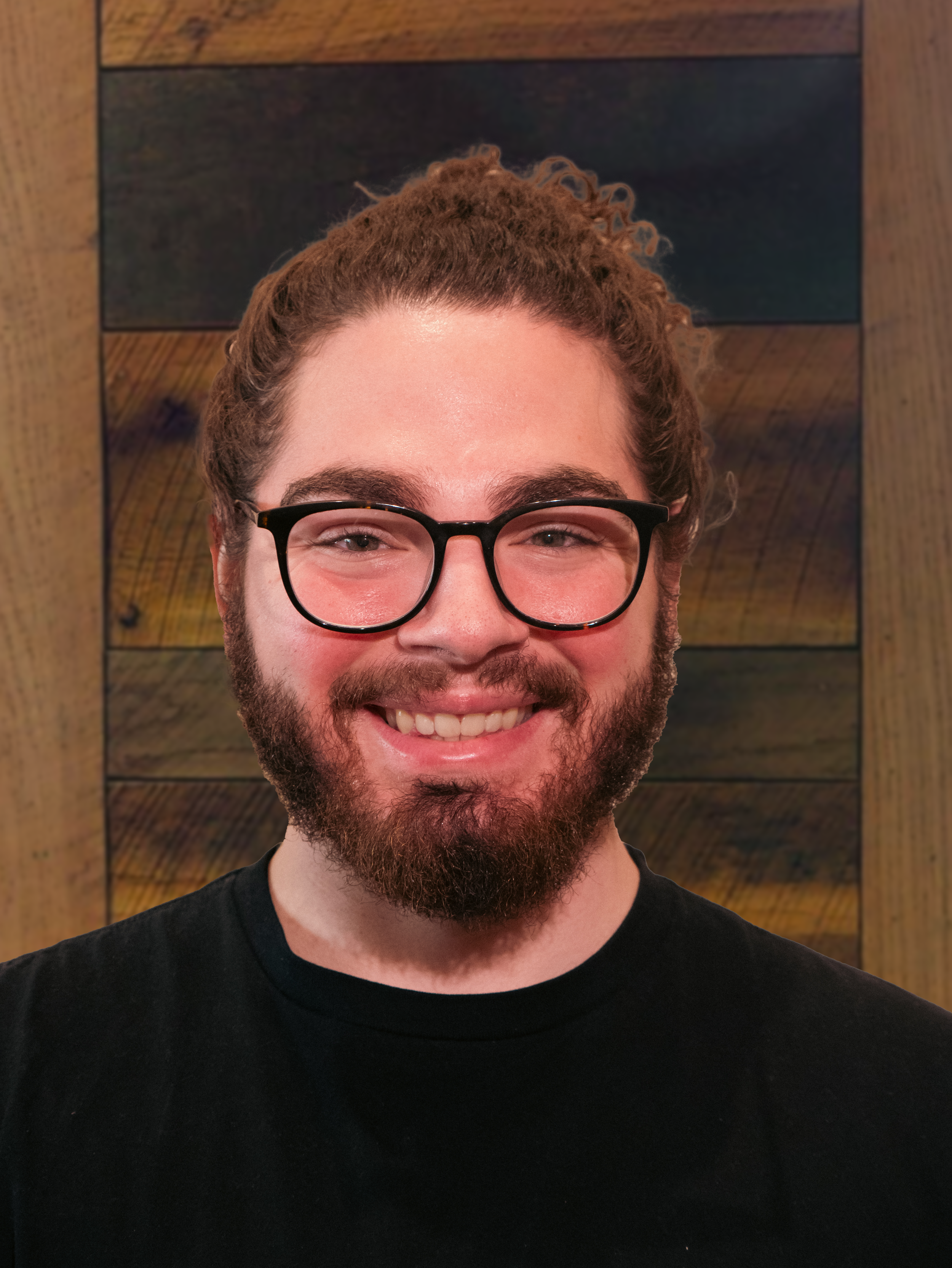 A smiling man with glasses, a beard, and curly hair against a wooden background.