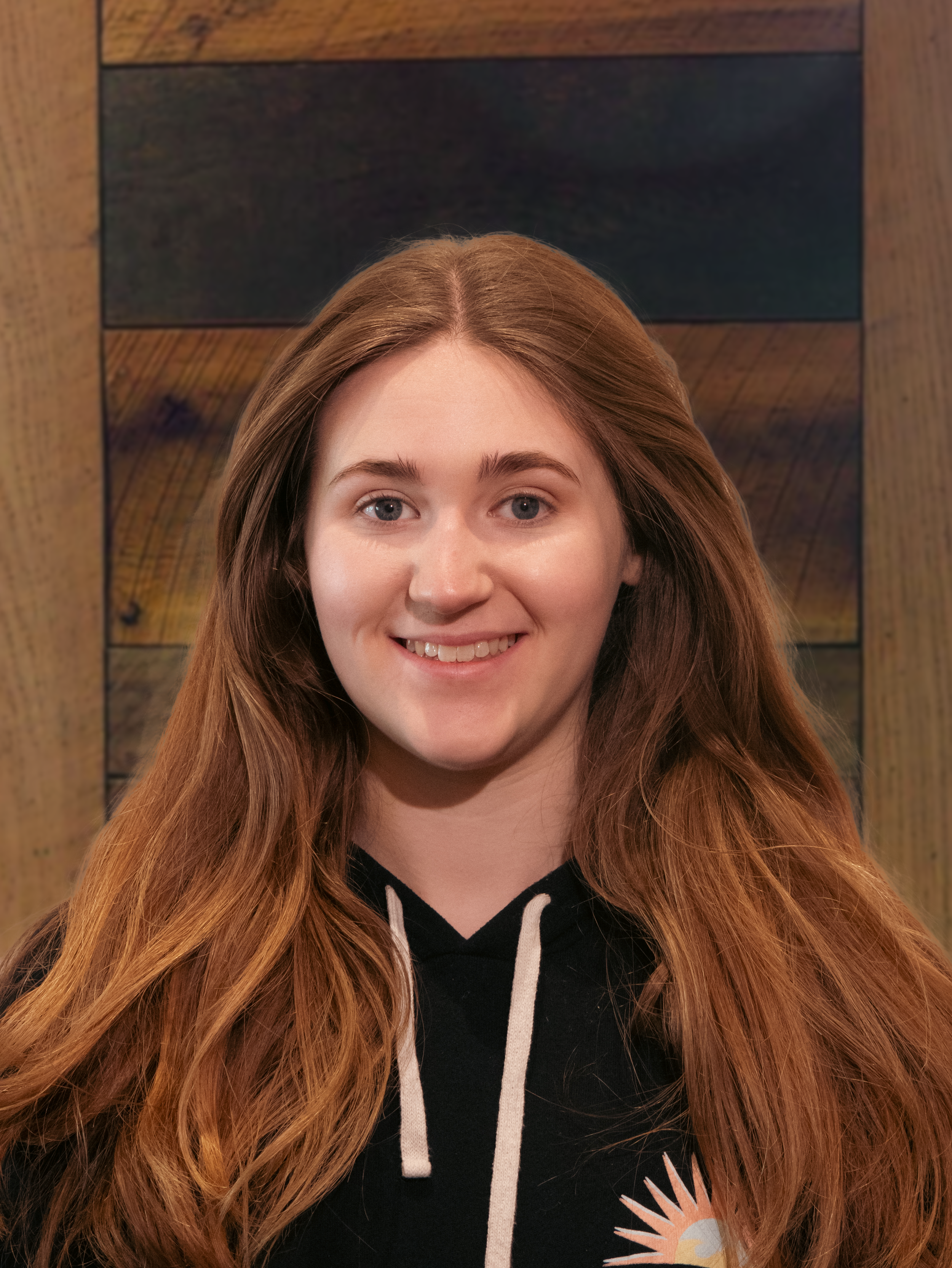 Portrait of a young woman with long red hair, smiling, wearing a black hoodie with a sun graphic, standing in front of a wooden wall.