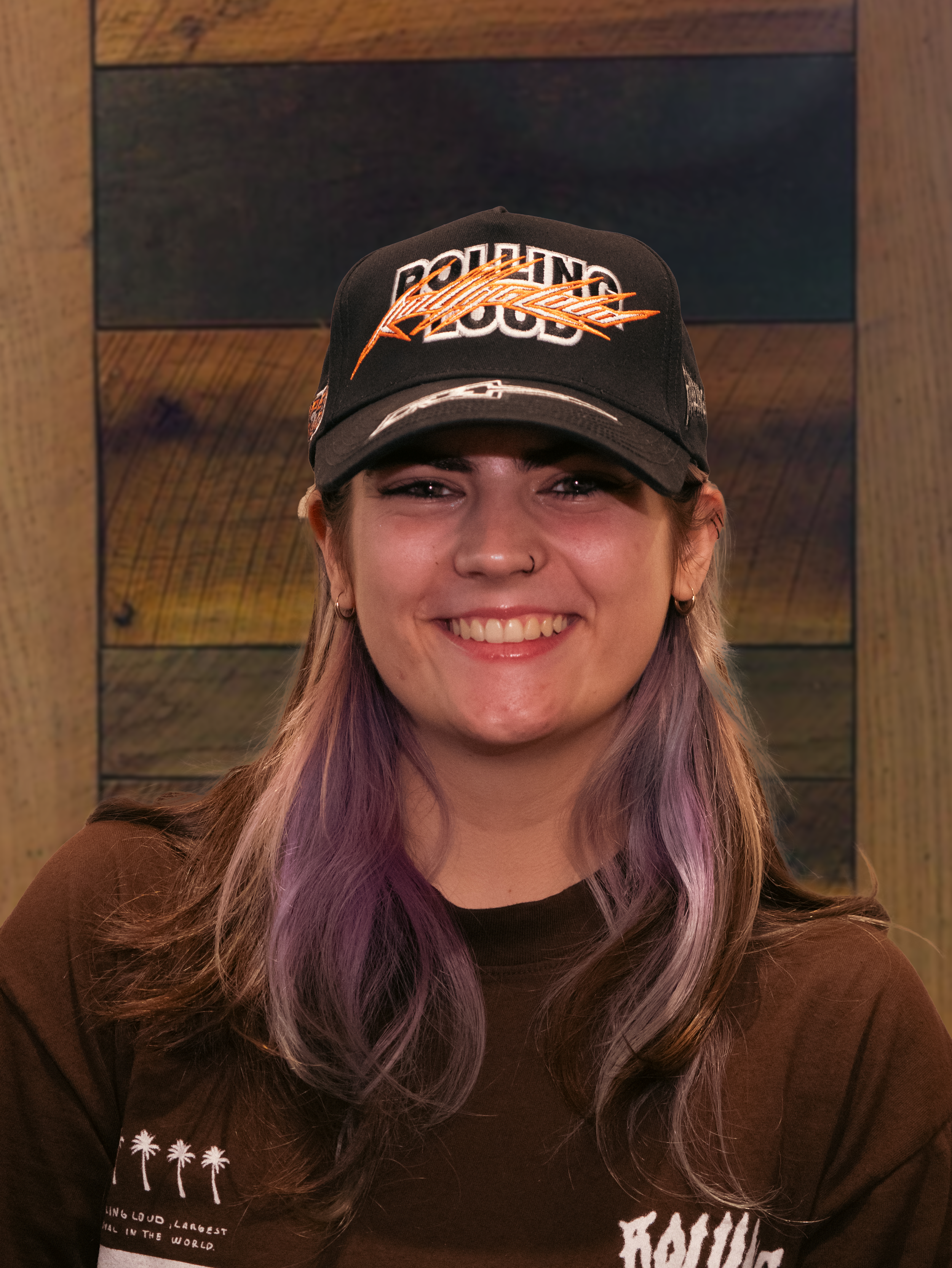 A young woman is smiling and wearing a black cap with 'Rolling Loud' and 'Radio' written on it, along with a brown shirt with palm trees and text on it. She has purple and brown hair and is standing against a wooden background.