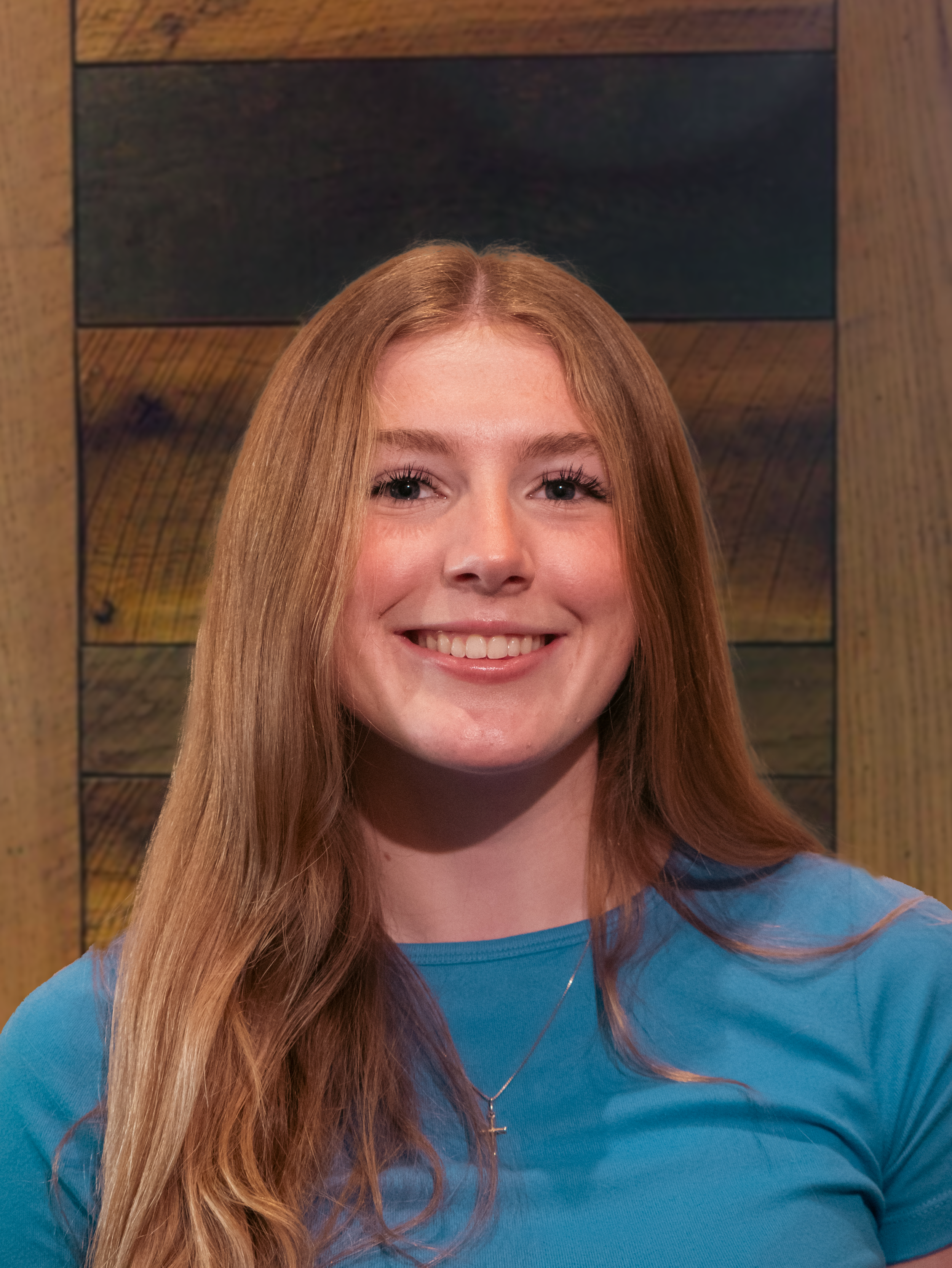Young woman with long red hair smiling, wearing a blue top and a small cross necklace, standing in front of wood-paneled wall.