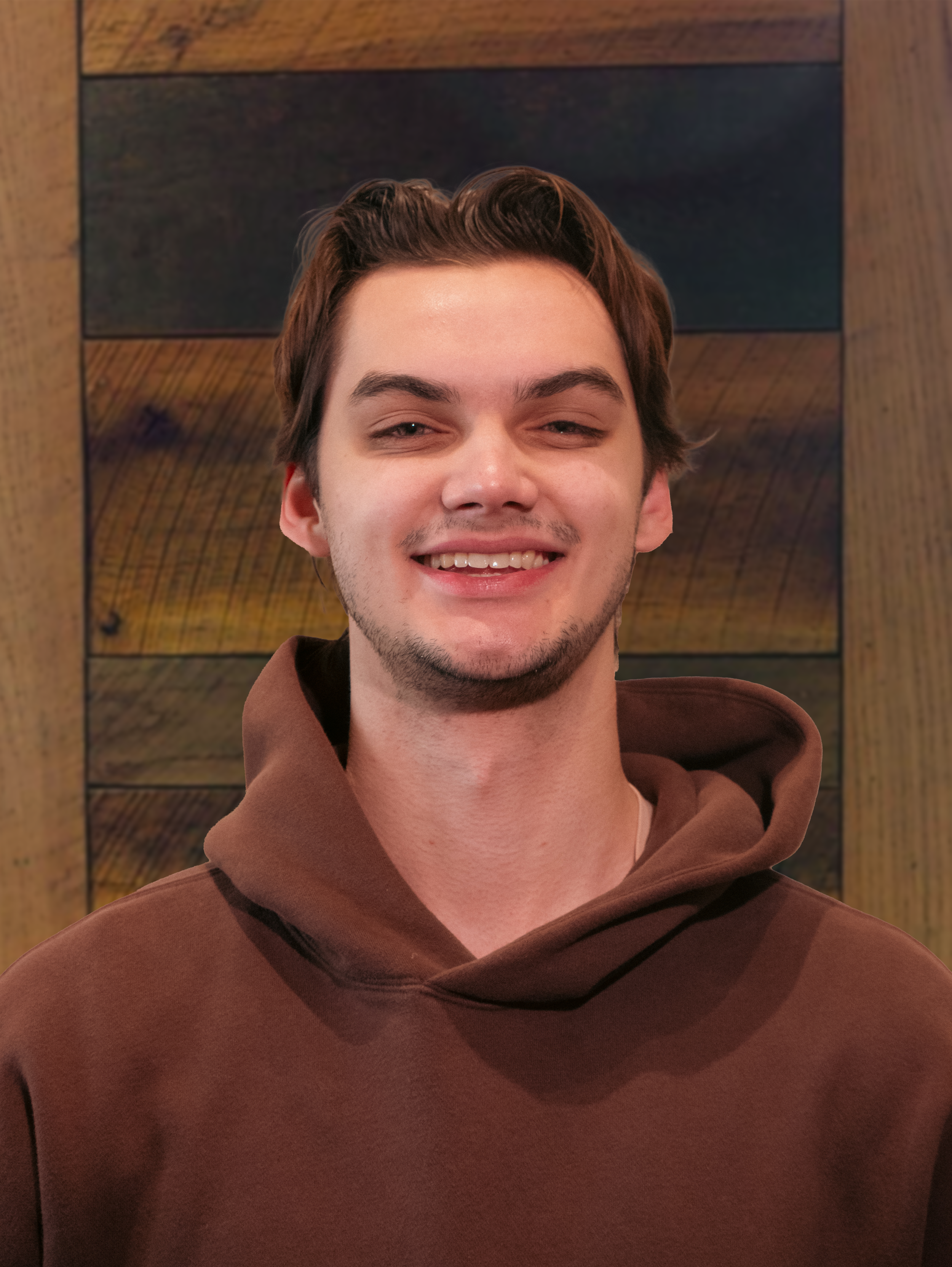 A young man with brown hair, light skin, and a short beard, smiling, wearing a brown hoodie, standing in front of a wooden wall.