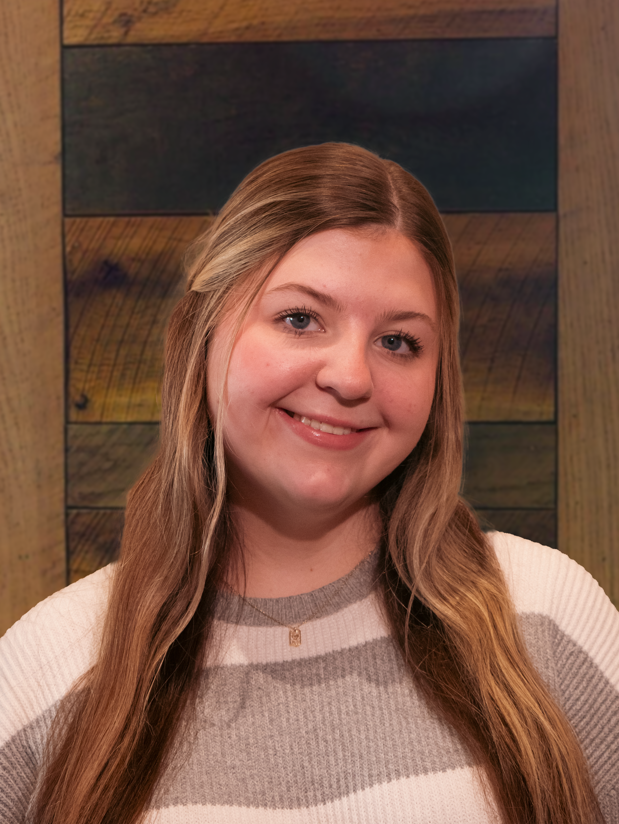 A young woman with long, wavy blonde hair smiling at the camera, wearing a striped sweater and a delicate necklace, standing in front of a wooden wall.