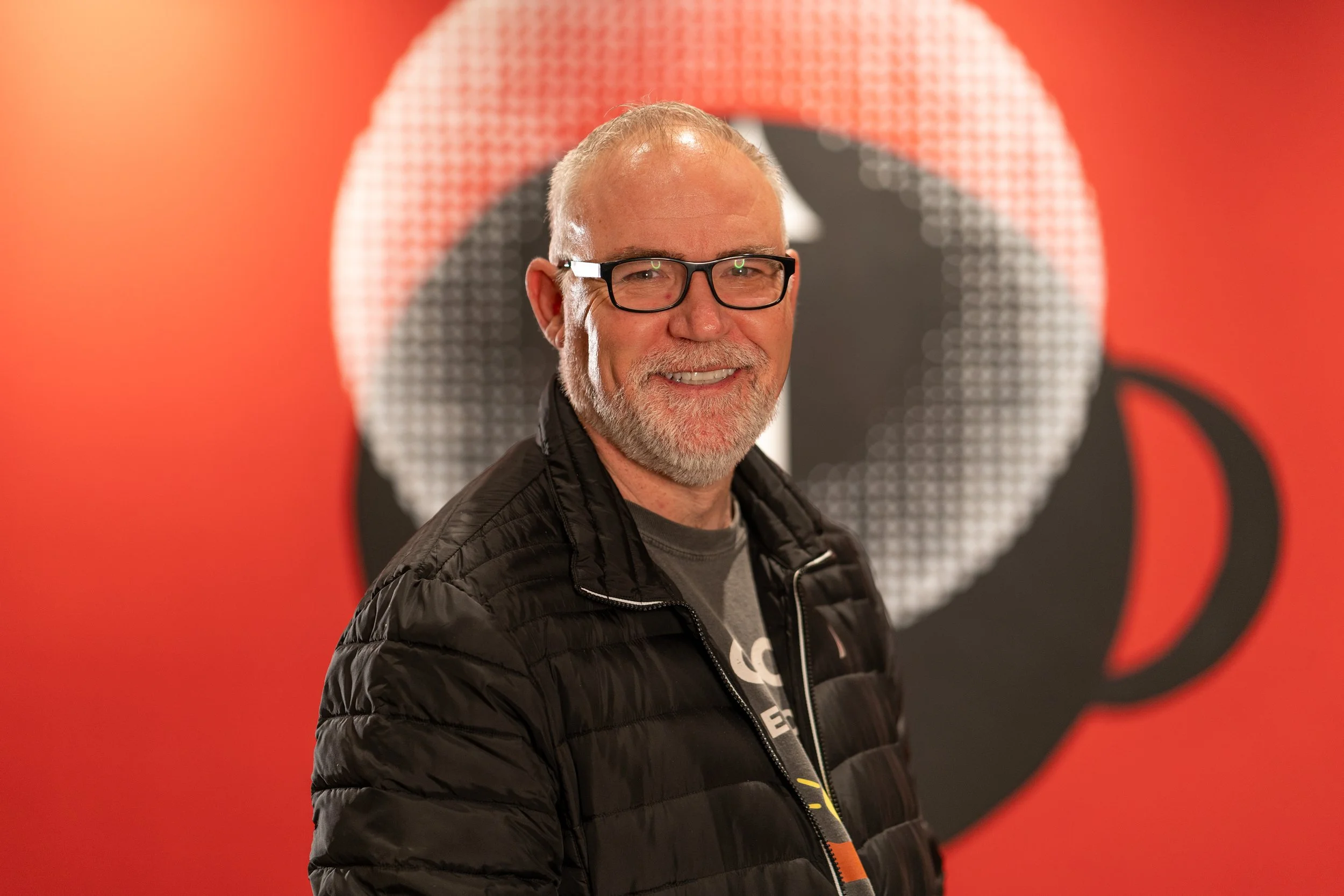 Smiling man with glasses and a beard, wearing a black jacket, standing in front of a red background with a Play Video button icon.