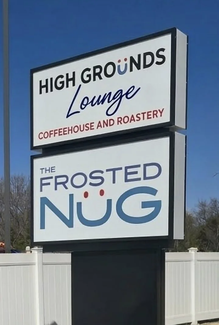 A sign for High Grounds Lounge, a coffeehouse and roastery, and The Frosted NüG, with stylized text and smiley face design in the logo.