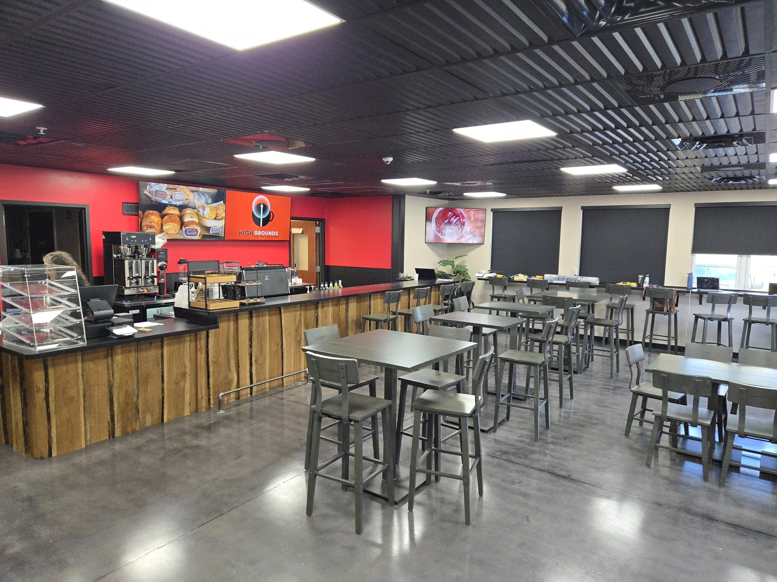 Empty cafe or coffee shop interior with wooden counter, tables, chairs, and digital screens on the walls, some with food images and branding.