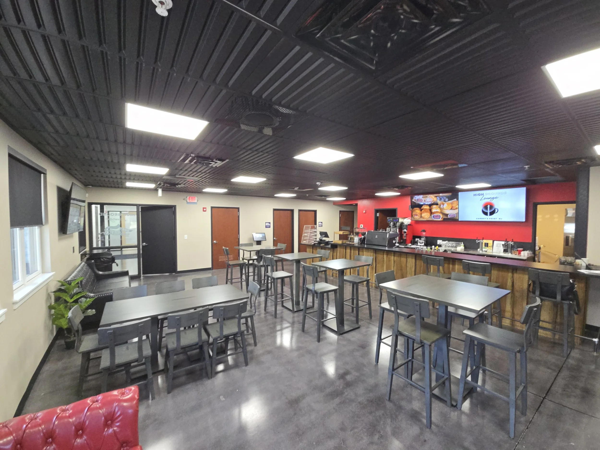 Interior of a modern fast food restaurant with black ceiling, gray walls, and a red accent wall behind the counter. The space has several black chairs and tables, a bar area with high stools, and a digital menu display showing images of burgers and fries.