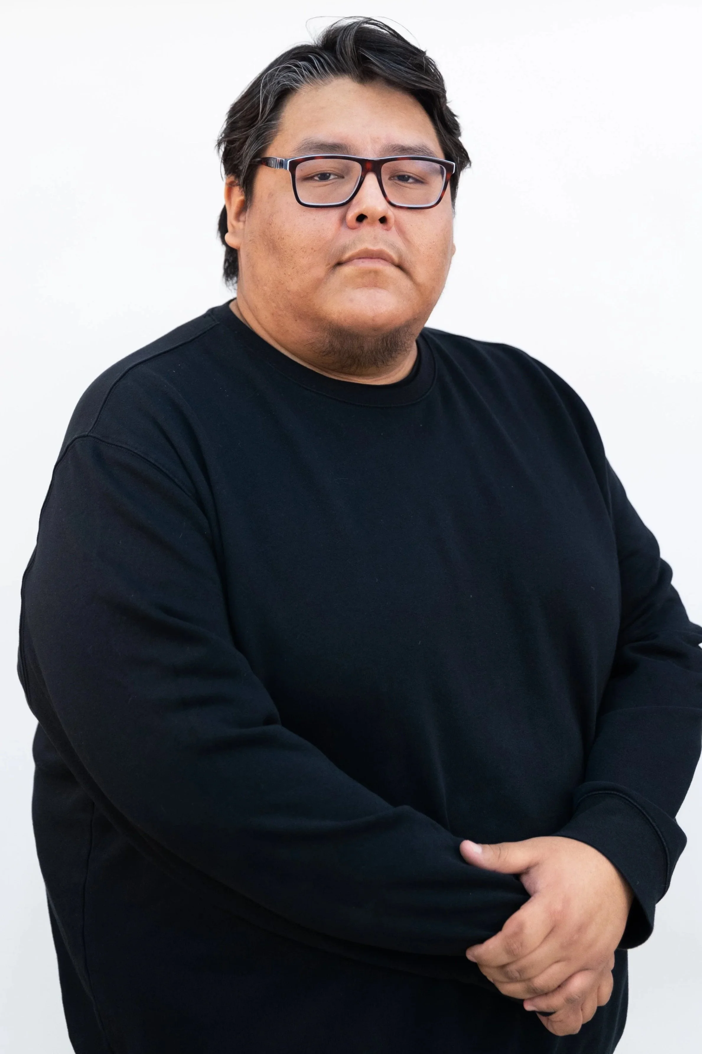 A man with black hair, glasses, and a beard, wearing a black long-sleeve shirt, stands against a white background with his arms crossed.