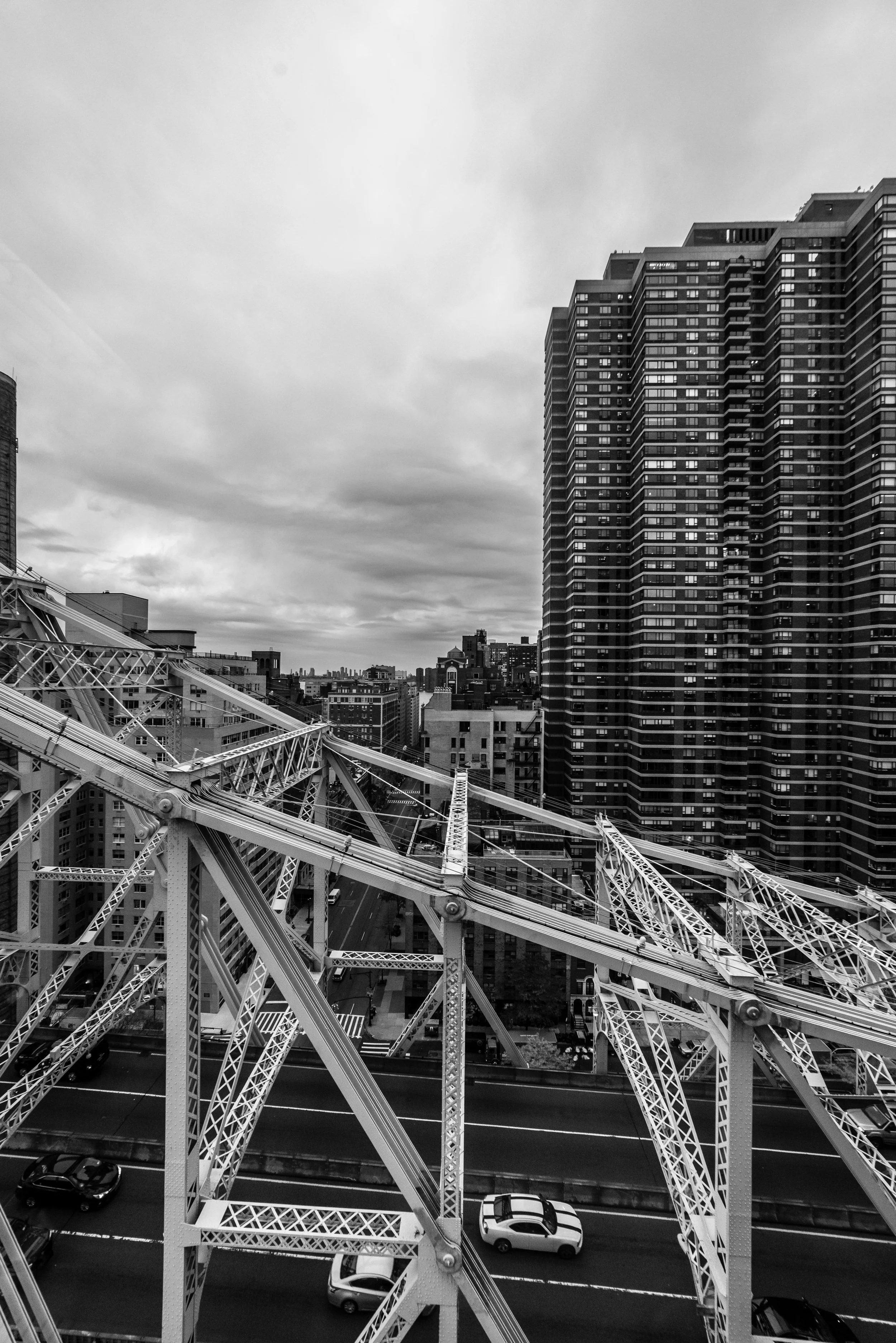 Black and white photo of an urban cityscape with tall modern buildings, a steel bridge structure in the foreground, and a cloudy sky.