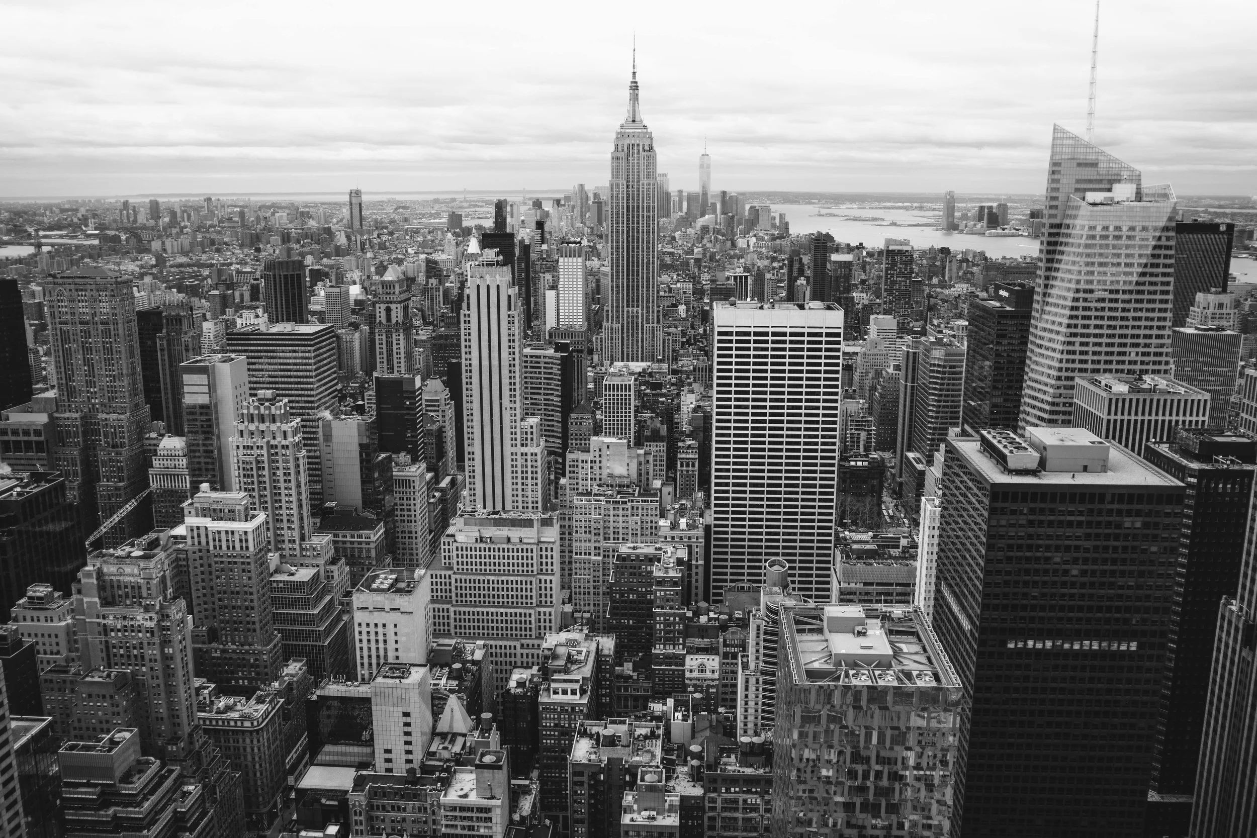 Black and white aerial view of New York City skyline with Empire State Building and other skyscrapers under cloudy sky.