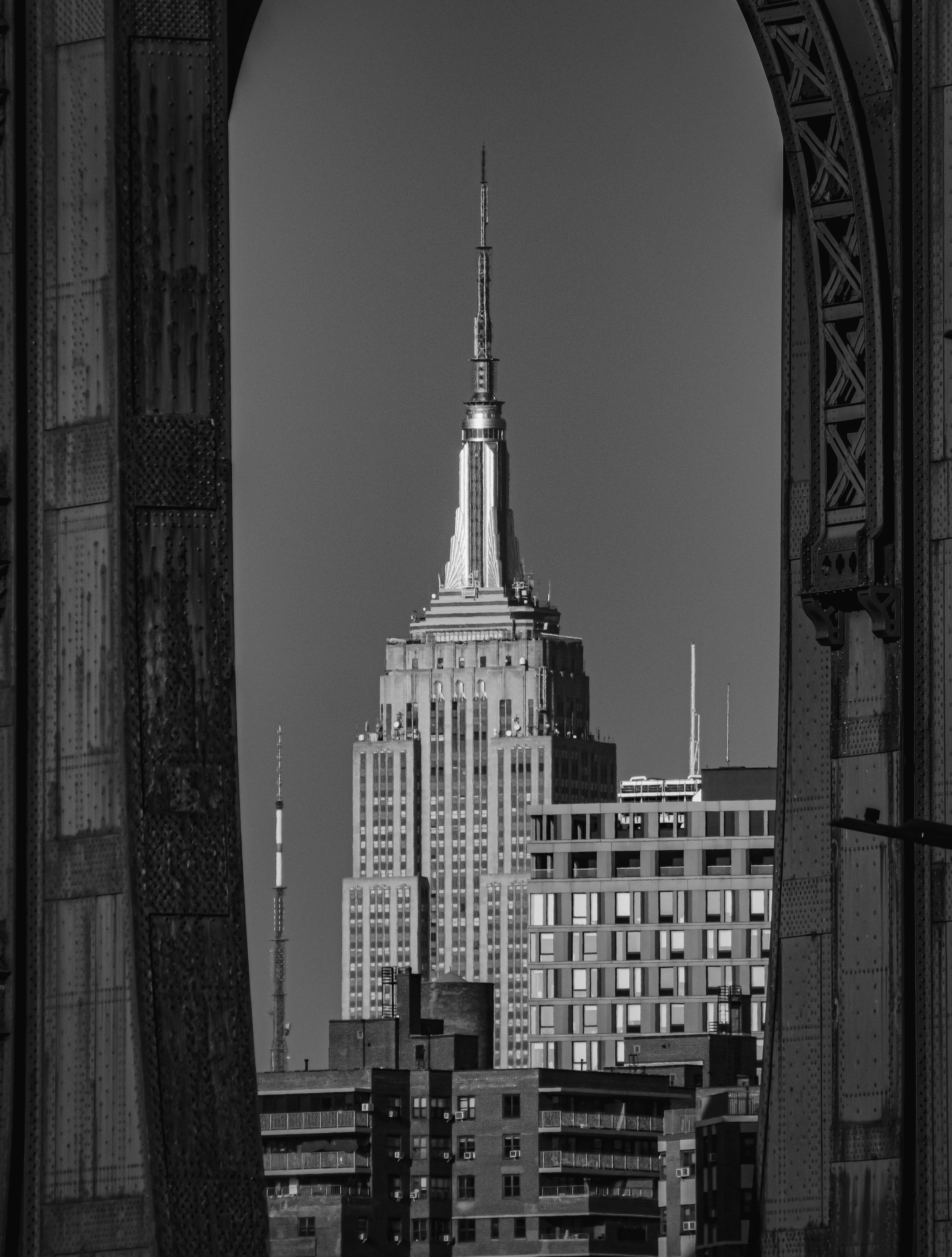 Black and white photo of the Empire State Building seen through a frame of industrial metal structures.