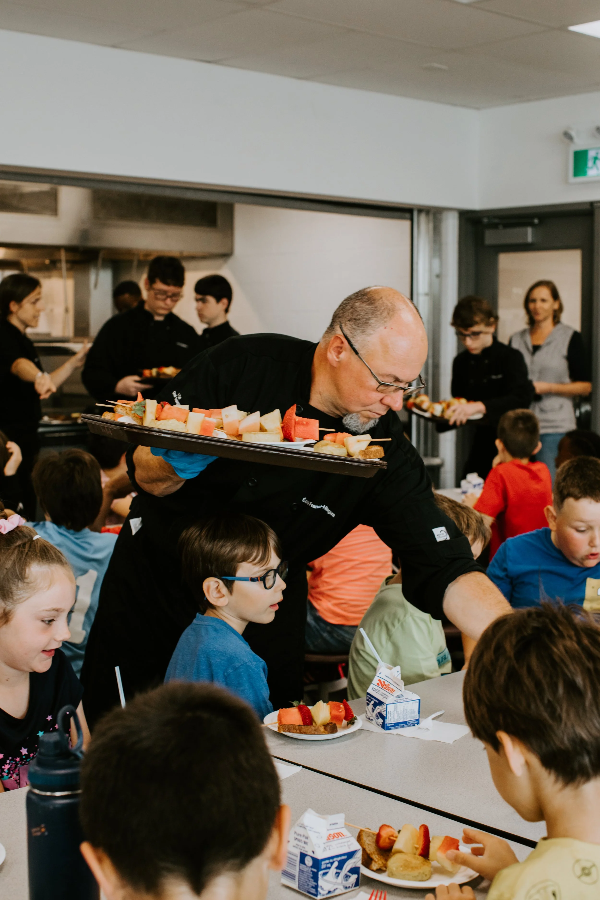 A group of children sitting at a cafeteria table with plates of sliced fruit and bread, and a man serving food. In the background, a few teenagers and an adult woman are waiting in line. The setting appears to be a school cafeteria.