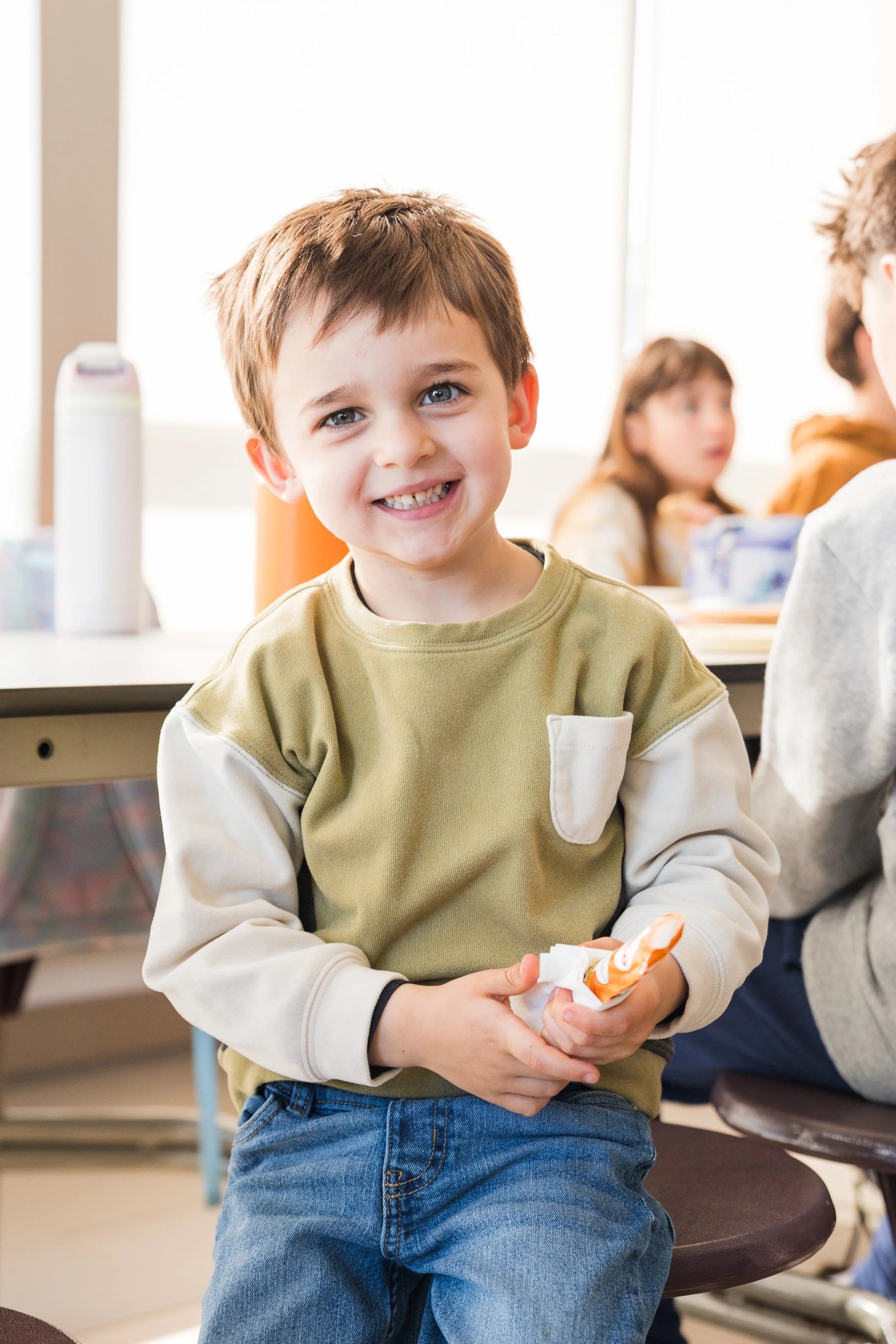 A young boy with brown hair and blue eyes smiling while holding a snack in his hands in a classroom setting with other children in the background.