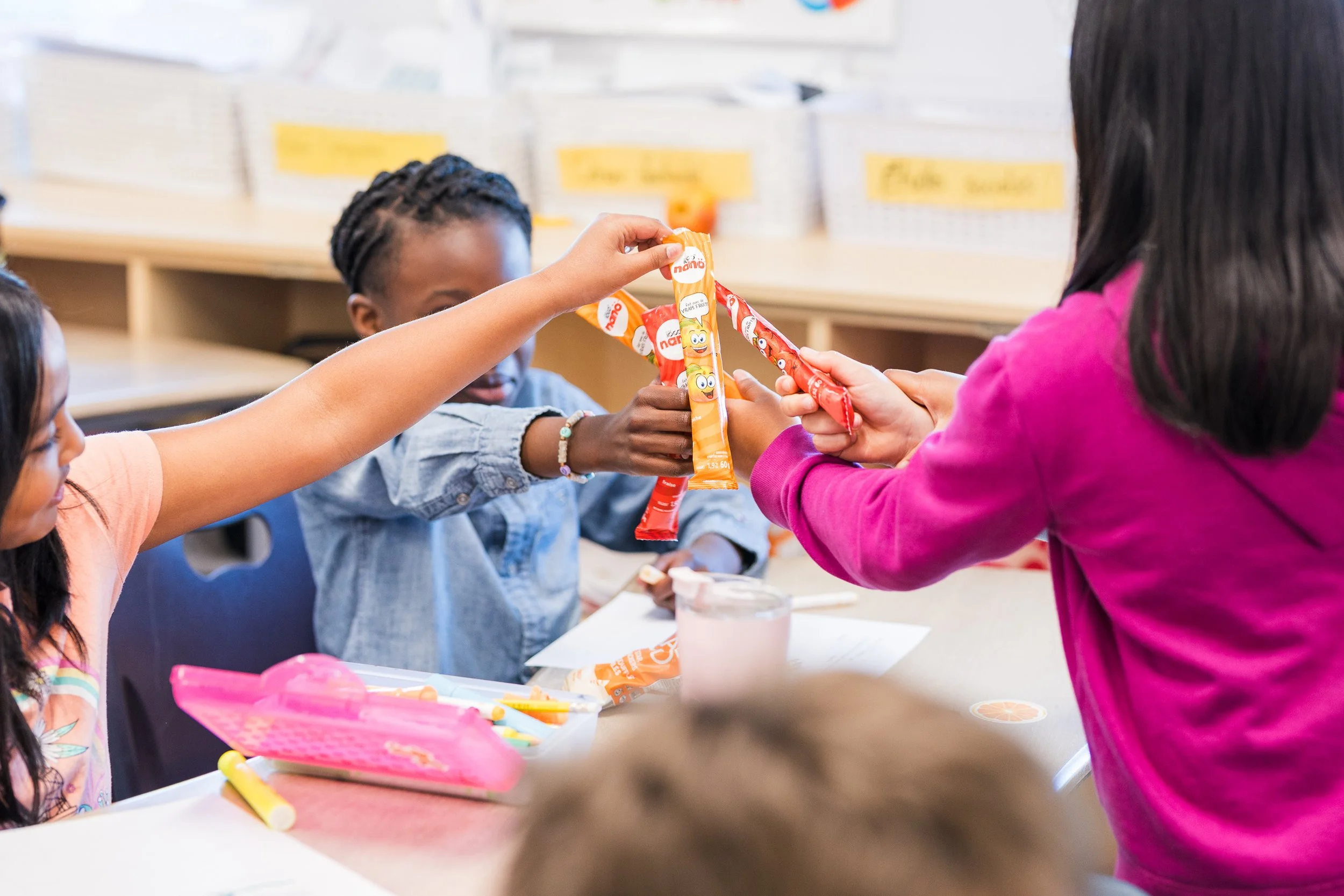 Children sharing orange snack packs in a classroom, with some snacks on the table and a few colorful objects nearby.