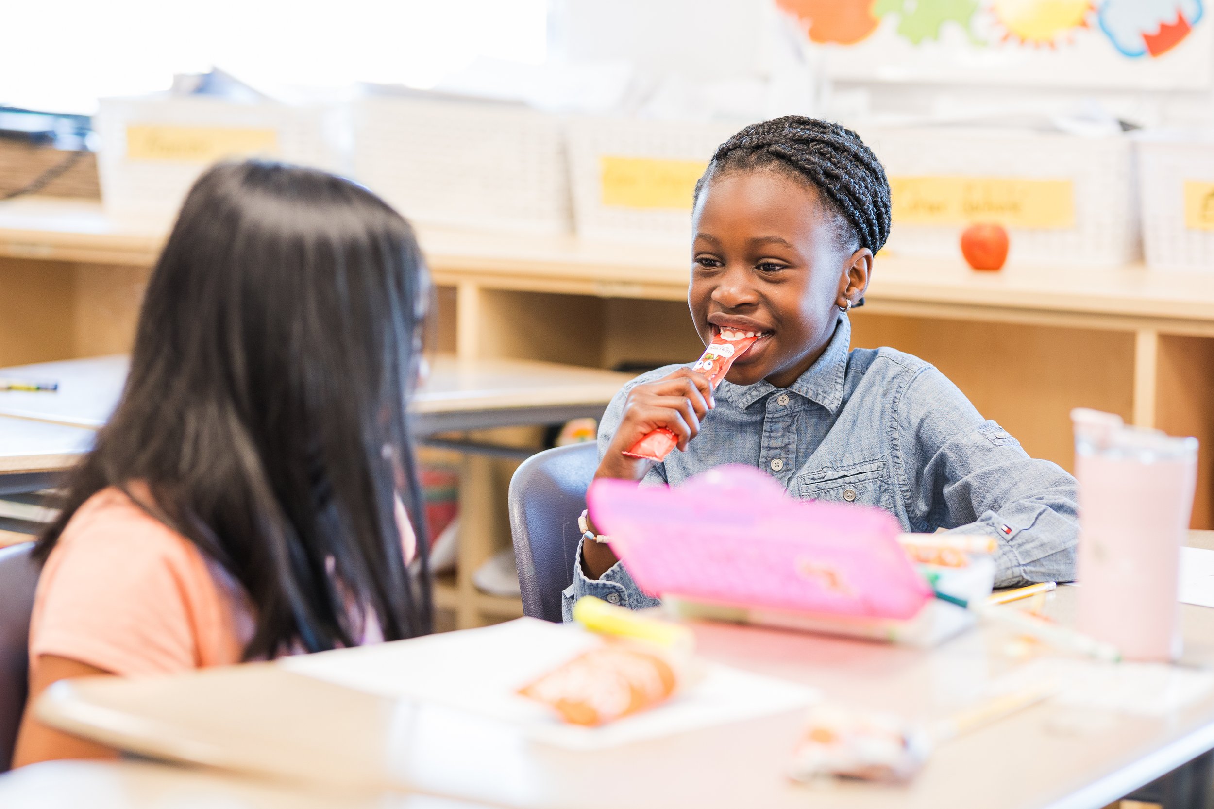 A young girl with braided hair smiling and holding a snack in a classroom setting with other children and colorful decorations.