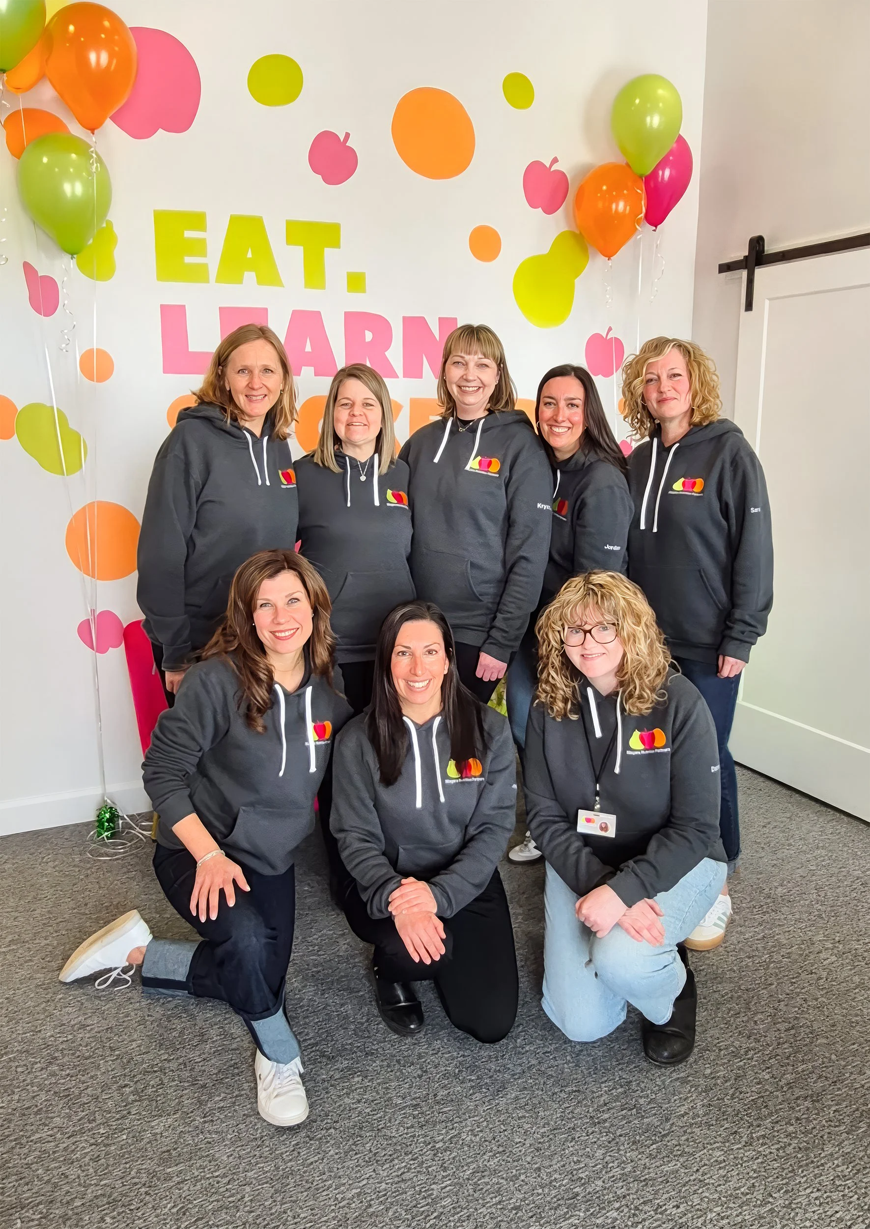 Group of eight women wearing matching dark hoodies with a logo, posing together in front of a decorative wall with balloons and a sign that says 'EAT. LEARN. GROW.'