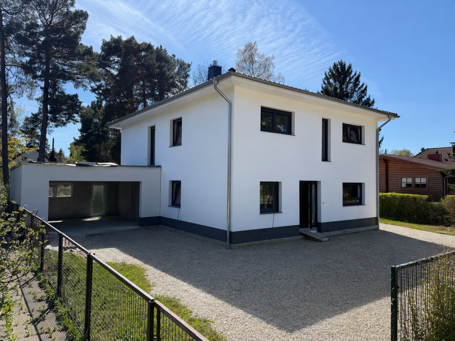 Modernes weißes zweistöckiges Haus mit Putzfassade und dunklen Fenstern, umgeben von Bäumen und einer Einfahrt aus Kies, bei sonnigem Himmel.