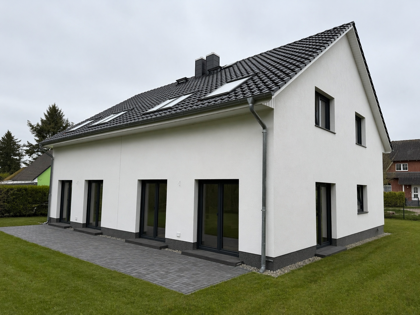 Modernes zweistöckiges, weißes Haus mit schwarzen Fenstern und Dach, Spazierweg am Garten, bei bewölktem Himmel.