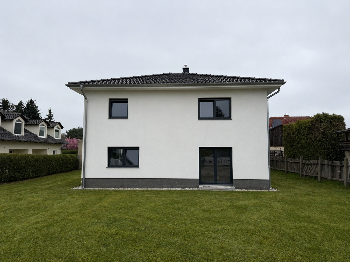 Ein weißes zweistöckiges Haus mit dunklem Dach und vier schwarzen Fenstern, umgeben von einem grünen Rasen und einem Holzzaun im Hintergrund.