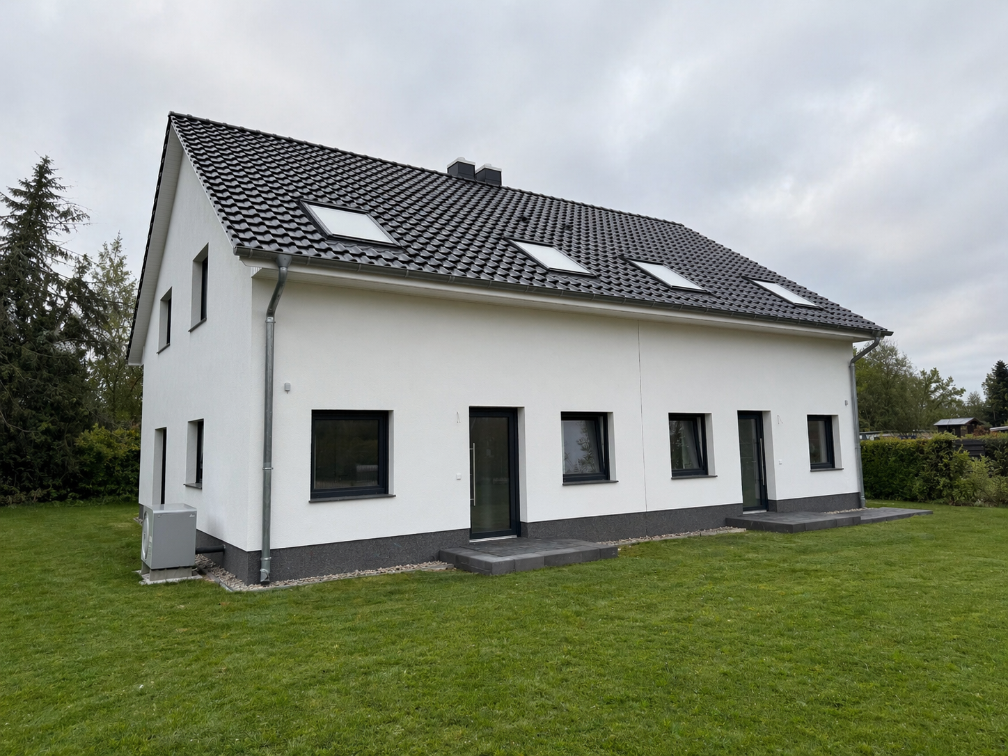 Ein modernes, zweistöckiges Einfamilienhaus mit weißer Fassade, schwarzen Fenstern und einem dunklen, schindelartigen Dach mit Dachfenstern, umgeben von einem gepflegten grünen Rasen.