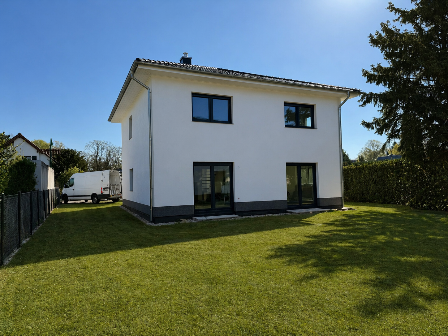 Ein modernes zweistöckiges Haus mit weißen Wänden und dunklen Fenstern, umgeben von einem gepflegten grünen Garten, auf einem sonnigen Tag.