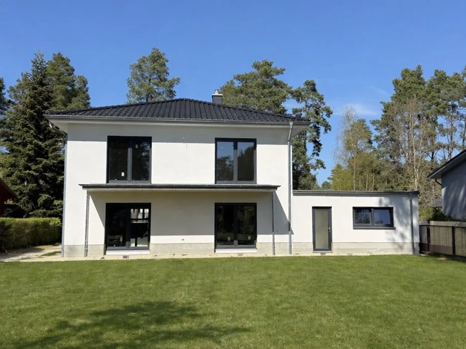 Ein modernes zweistöckiges Haus mit weißen Wänden, schwarzen Fenstern und einem dunklen Dach, umgeben von einem grünen Garten und Bäumen.