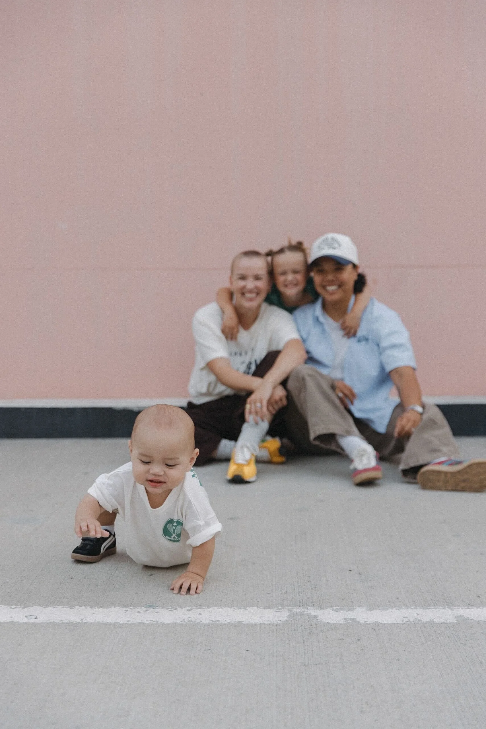 A young child crawling on a gray concrete floor with a pink wall in the background, while three adults sit behind him, smiling and hugging each other.