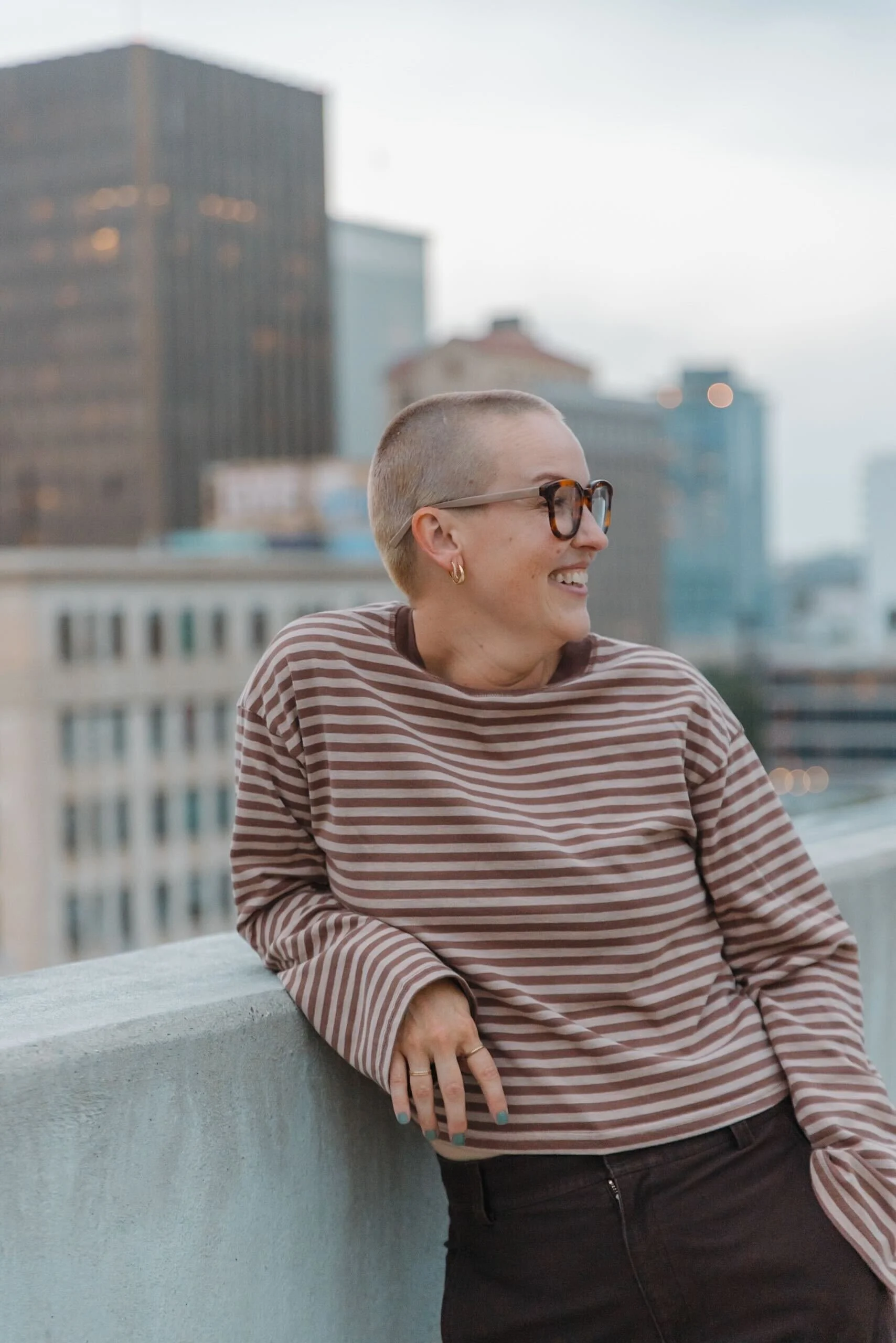A person with a shaved head and glasses leaning against a rooftop ledge, smiling with city buildings in the background.
