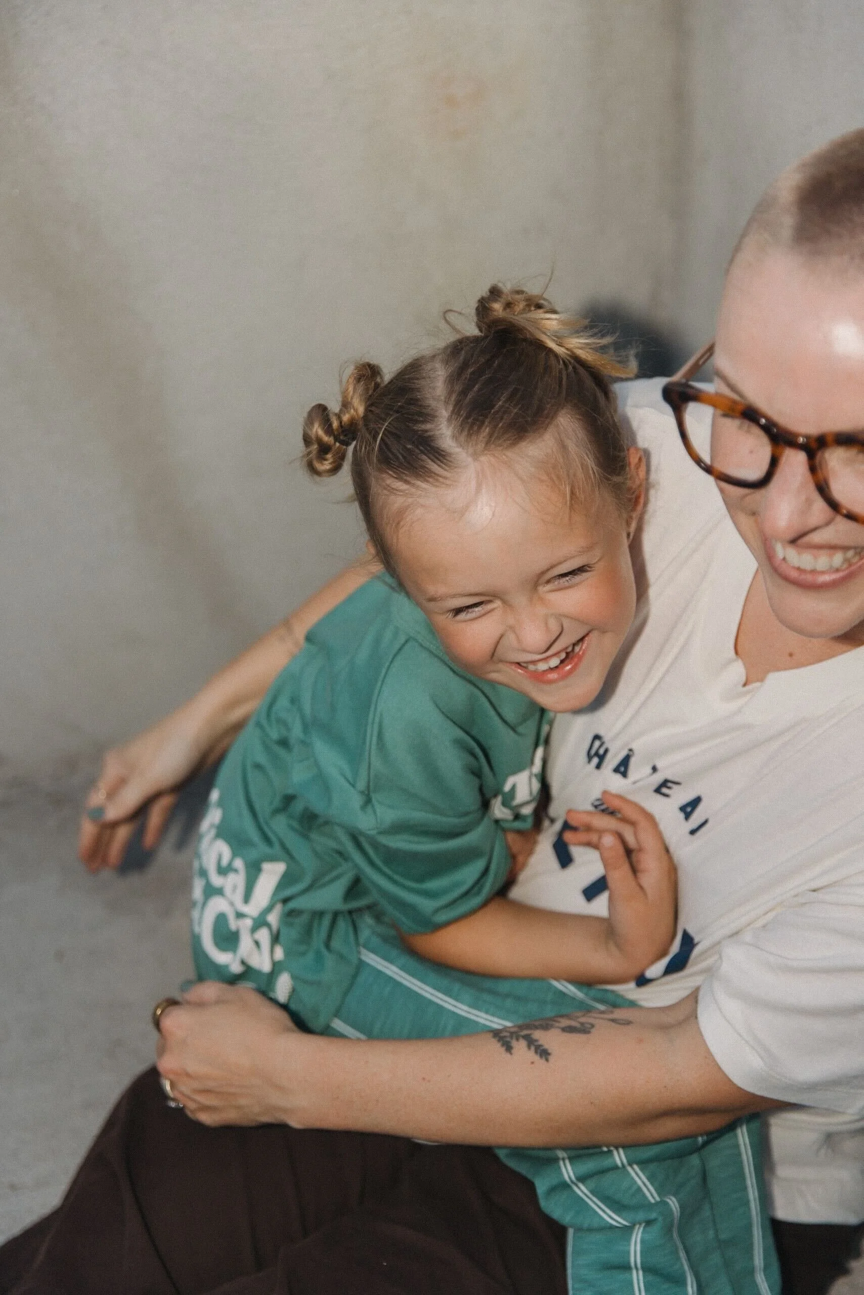 A young girl with brown hair in buns and a big smile hugging a woman with glasses, tattoo, and short hair. They are sharing a joyful moment indoors.