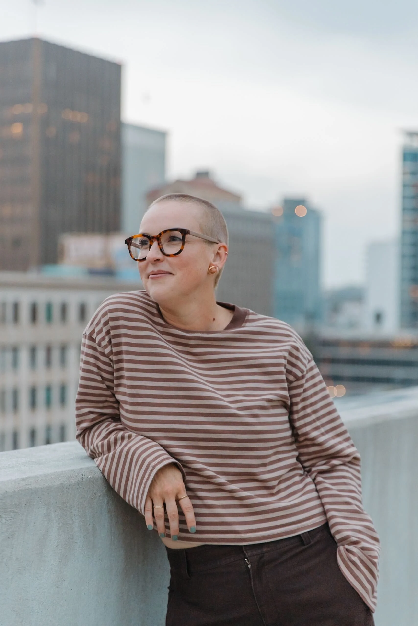 A woman with a shaved head wearing glasses, a striped brown and beige long-sleeve shirt, and dark pants, leaning on a rooftop ledge in an urban setting with tall buildings in the background.