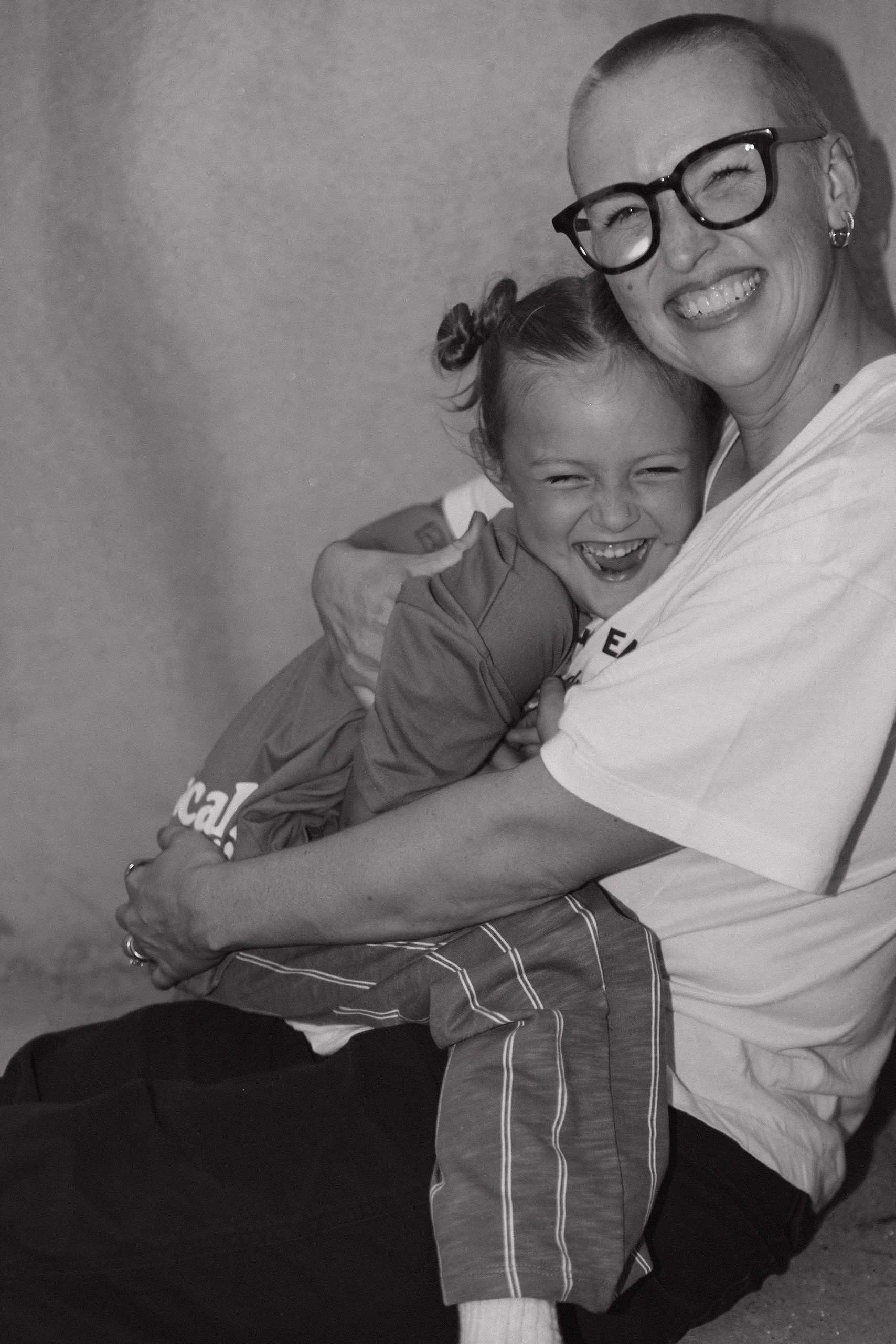A smiling woman with glasses and a shaved head is hugging a young girl with pigtails, both laughing happily.