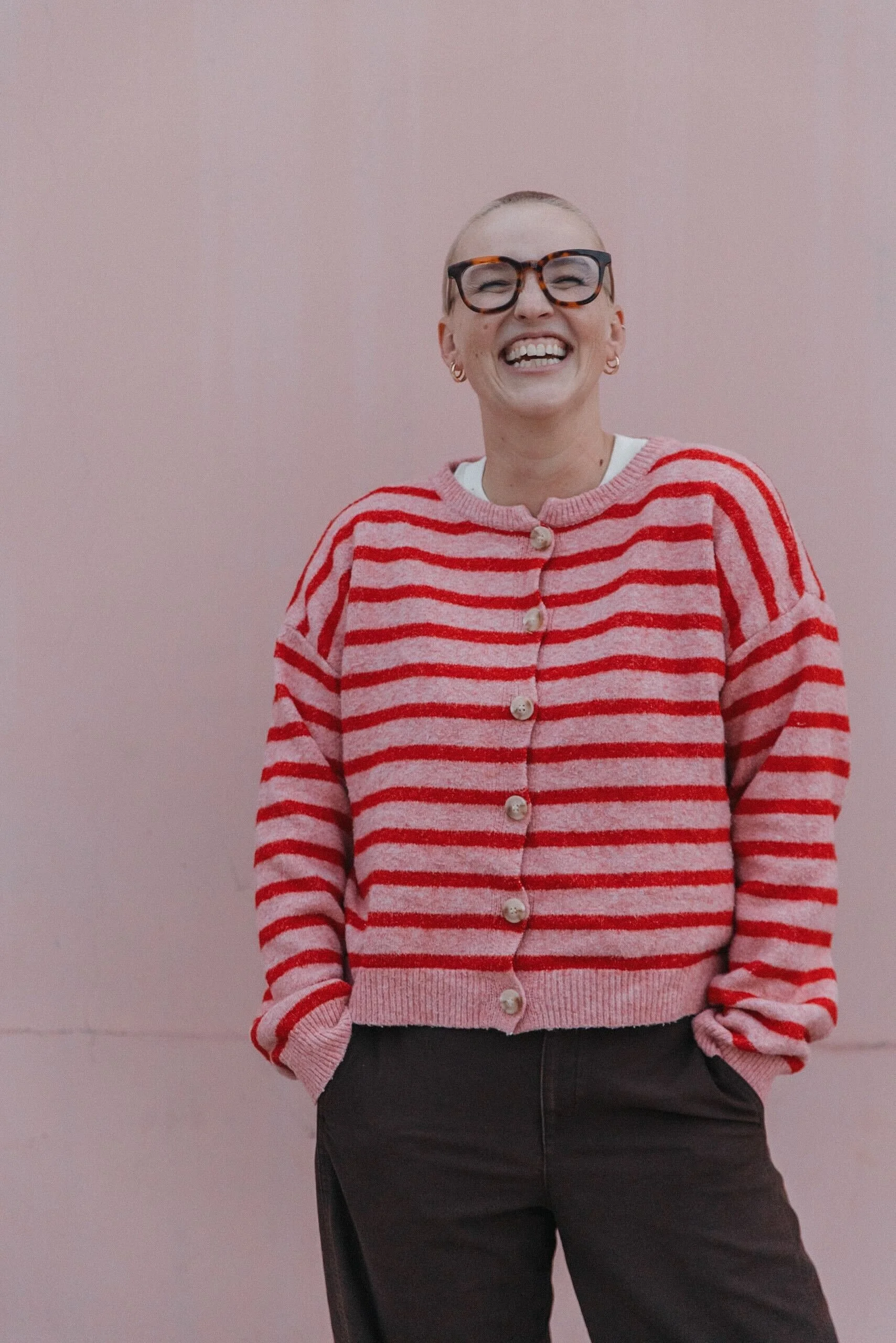 A woman with short hair, glasses, and earrings is smiling while wearing a pink and red striped cardigan and black pants against a pink wall.