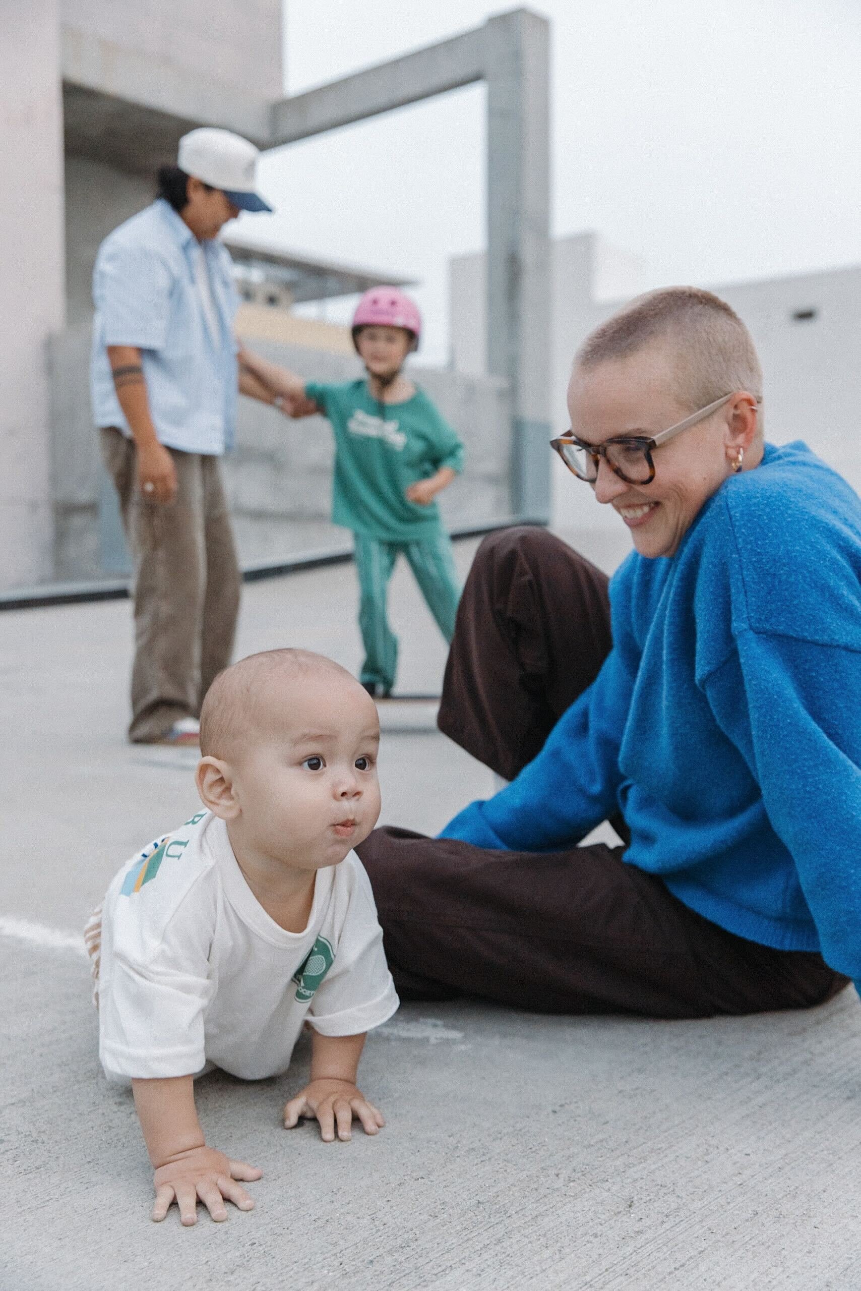 A baby crawling on the ground with an adult woman smiling nearby, while two children with helmets play in the background on a grey outdoor surface.