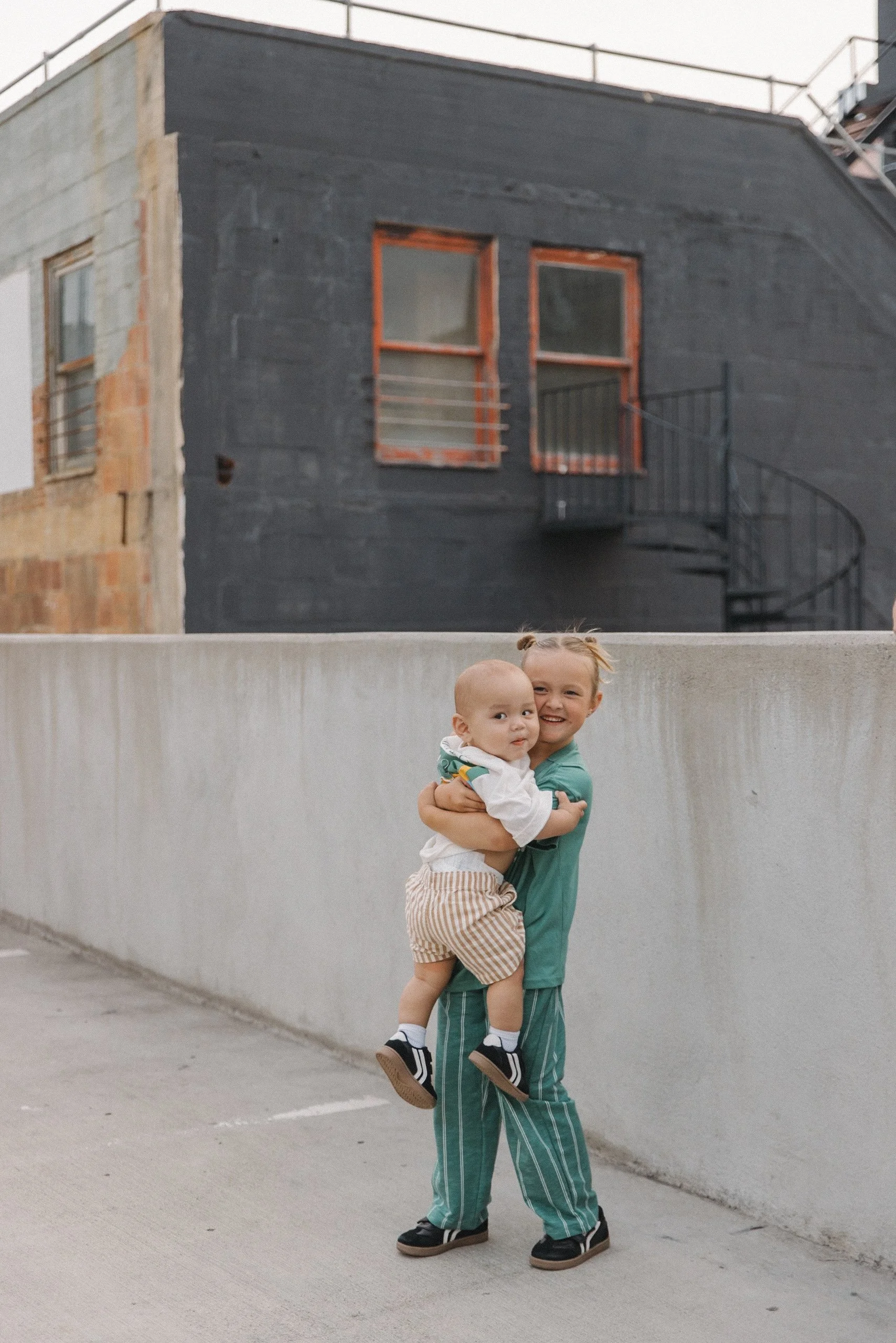 A young girl hugging a baby boy on a rooftop parking lot with an urban backdrop of a building with dark gray walls and orange window frames.