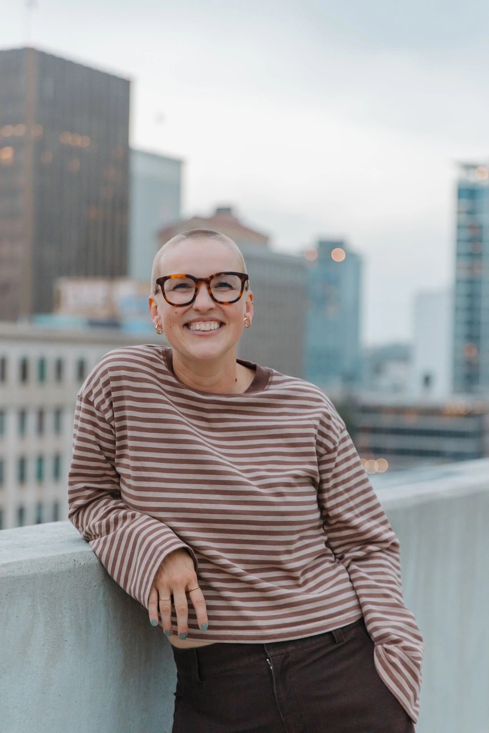 A smiling woman with short hair, glasses, and earrings, leaning on a rooftop ledge with city buildings in the background, during dusk.