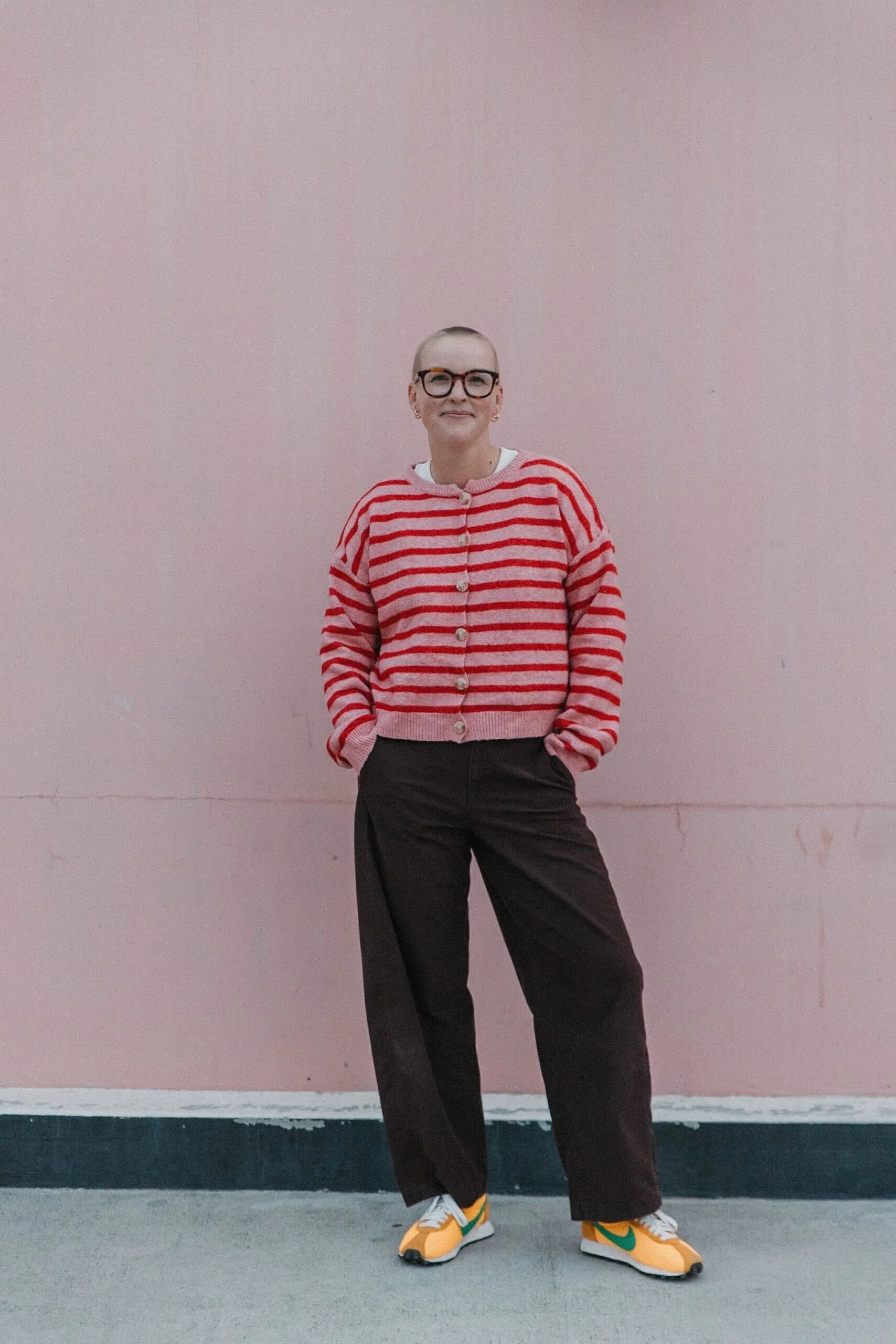 Person with short hair, glasses, wearing a red and pink striped cardigan, black pants, and yellow sneakers, standing against a plain pink wall.