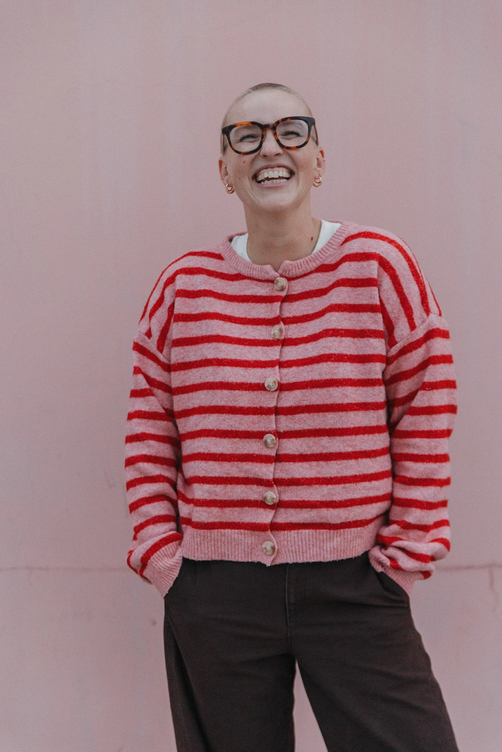 A smiling woman with short hair, glasses, earrings, wearing a pink and red striped sweater, standing against a pink wall.