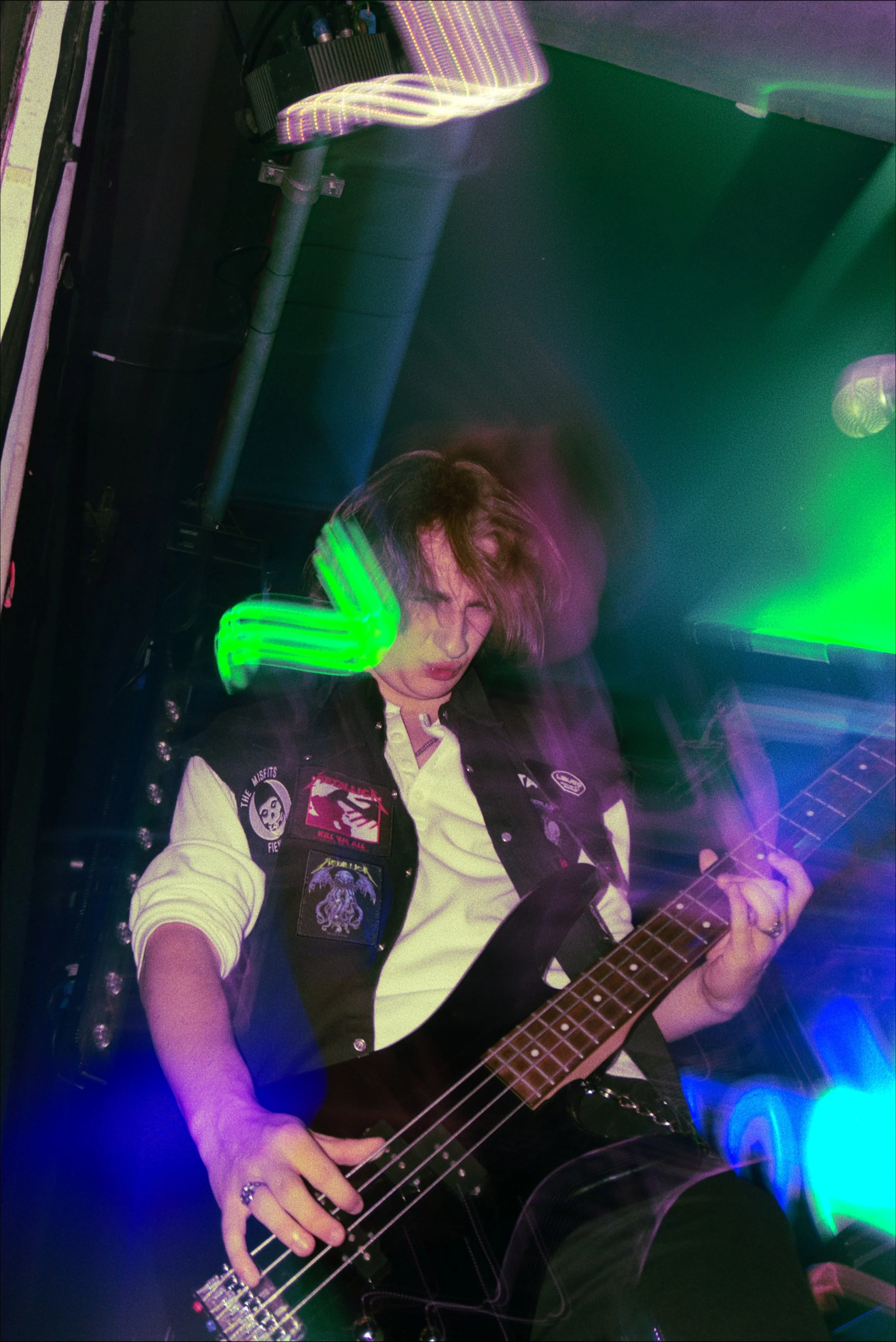 Person with shoulder-length hair playing a bass guitar in a room with colorful, moving lights.